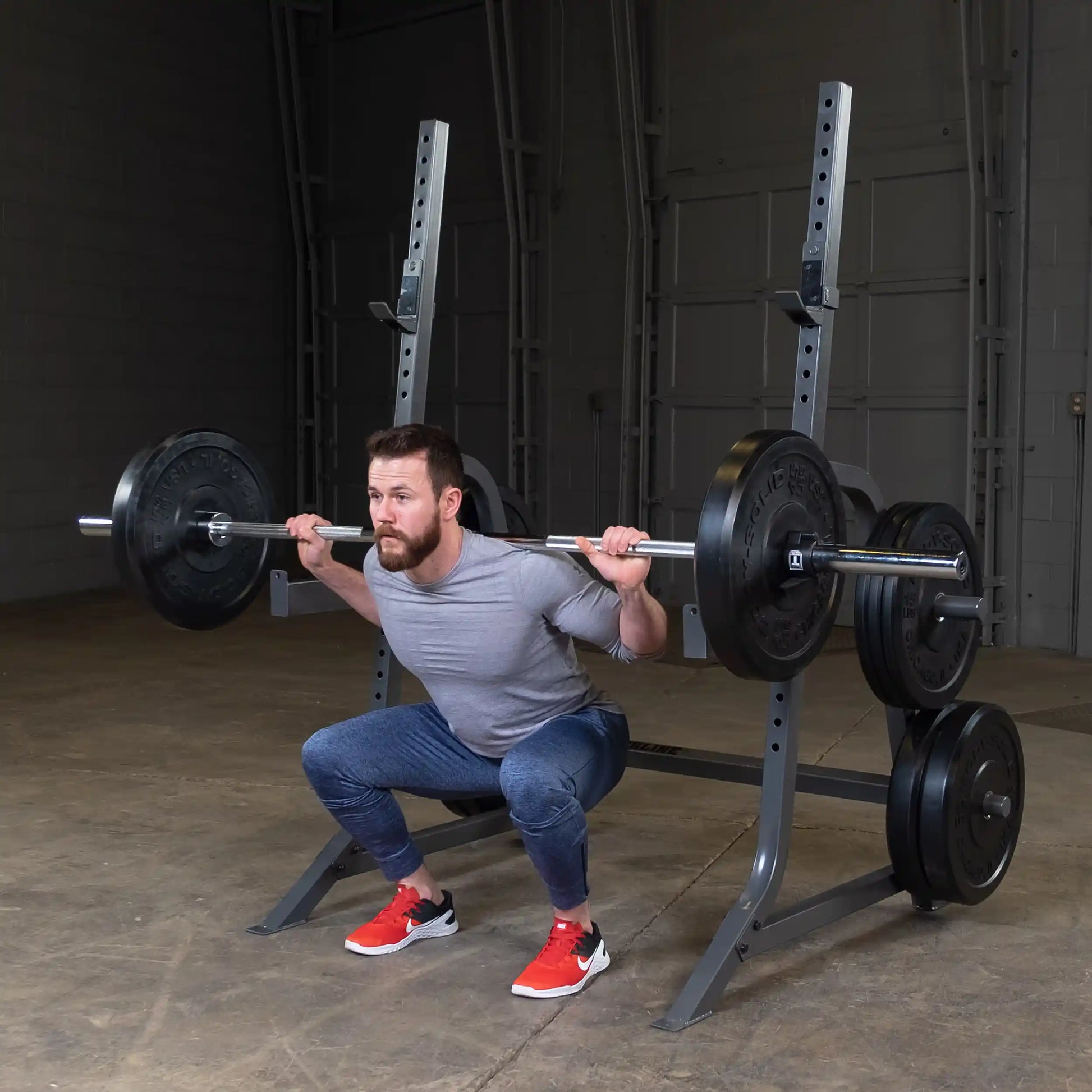 A man performing a barbell squat in a gym, wearing a gray shirt and blue pants, with red and black athletic shoes, in front of a squat rack loaded with weights.