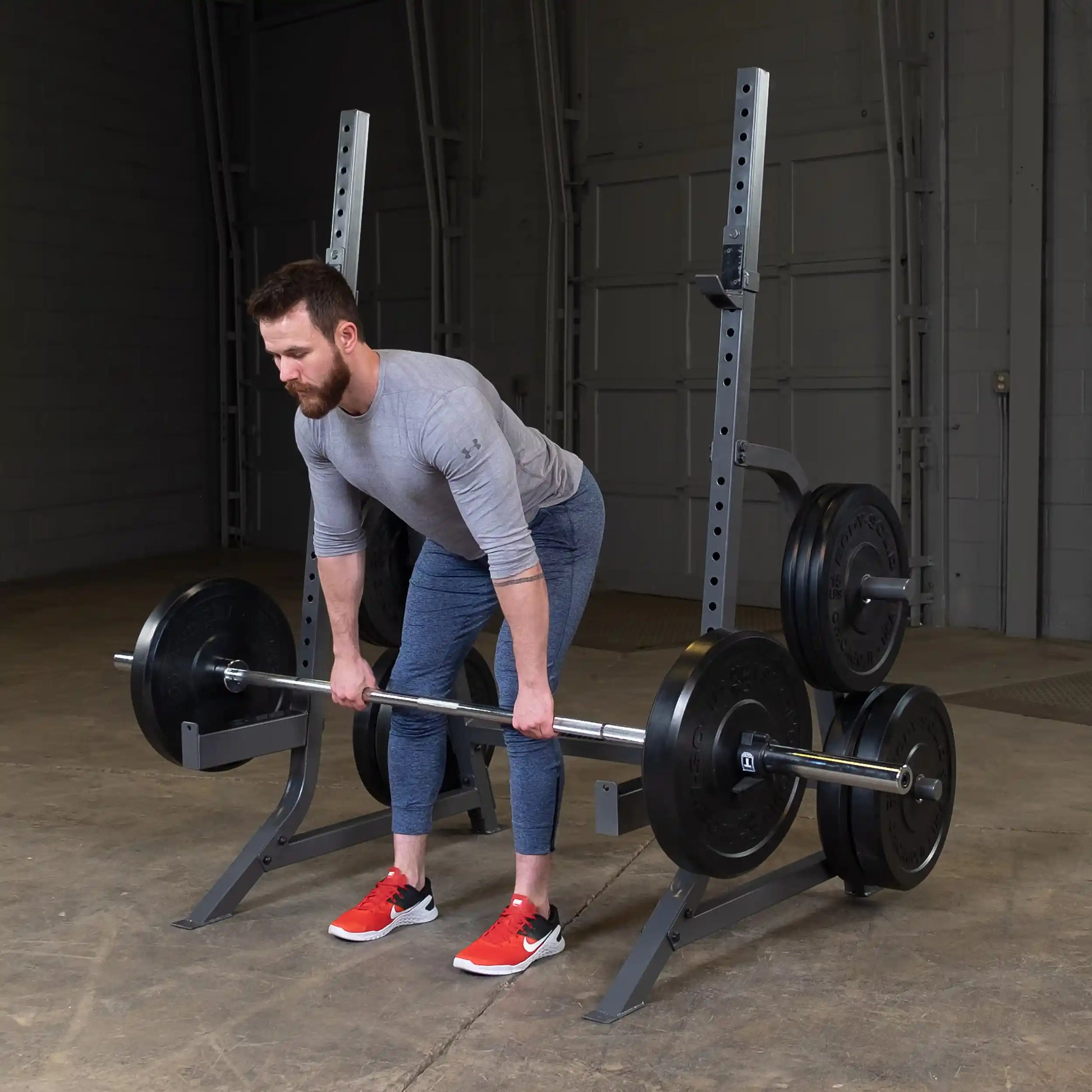A man in a gray long-sleeve shirt and blue athletic pants is performing a deadlift with a barbell loaded with weight plates in a gym setting. The background features gym equipment and concrete flooring.