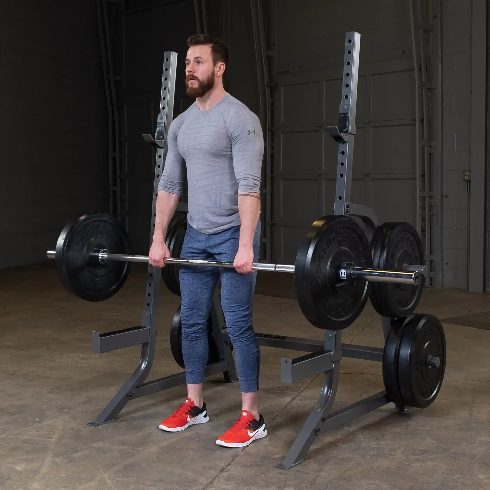 A man in a gray long-sleeve shirt and blue athletic pants stands in a gym, preparing to lift a barbell loaded with weight plates. He is wearing red and black athletic shoes and looks focused.