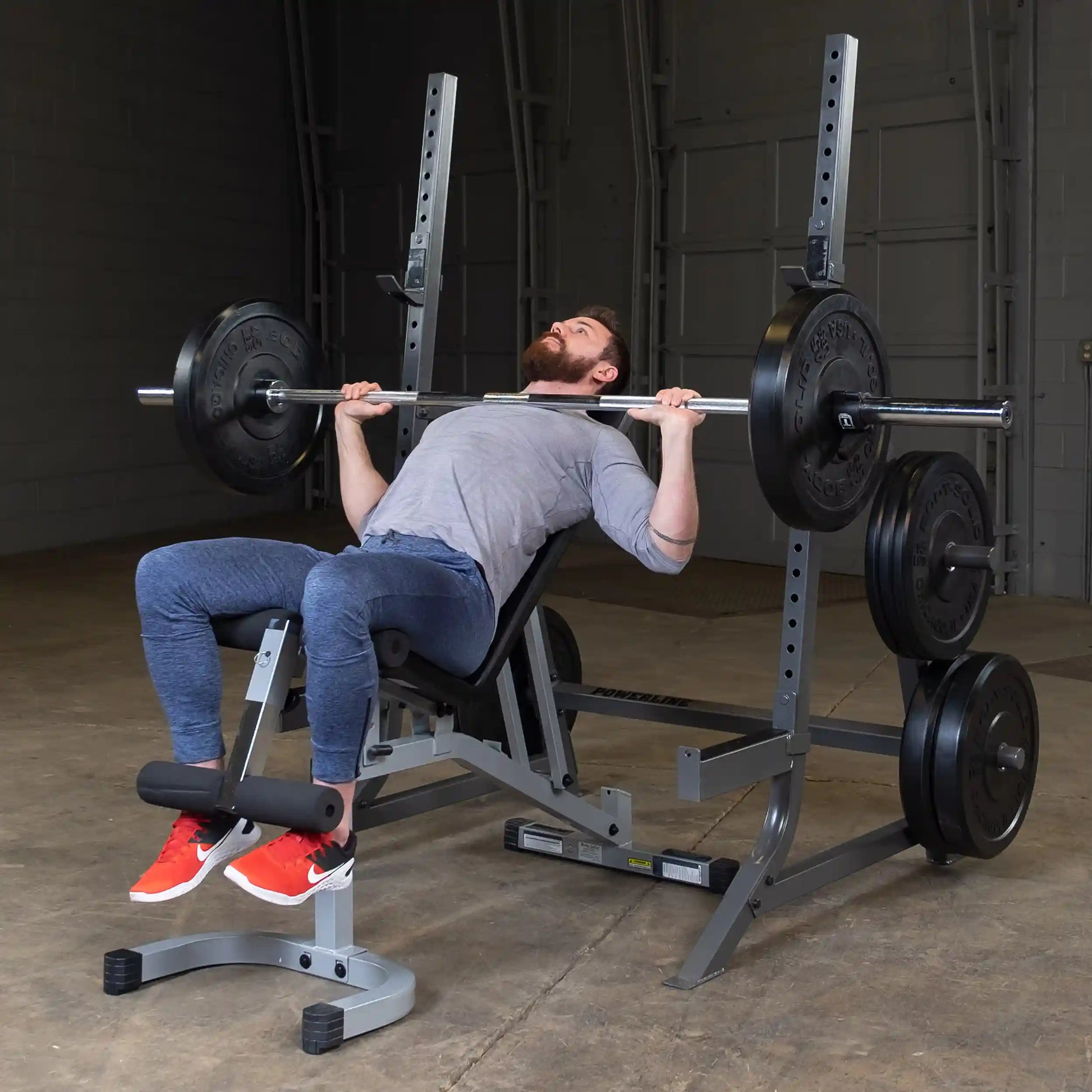 A man performing a bench press exercise with a barbell loaded with weights, seated on an adjustable bench in a gym setting, wearing a gray shirt and blue athletic pants.