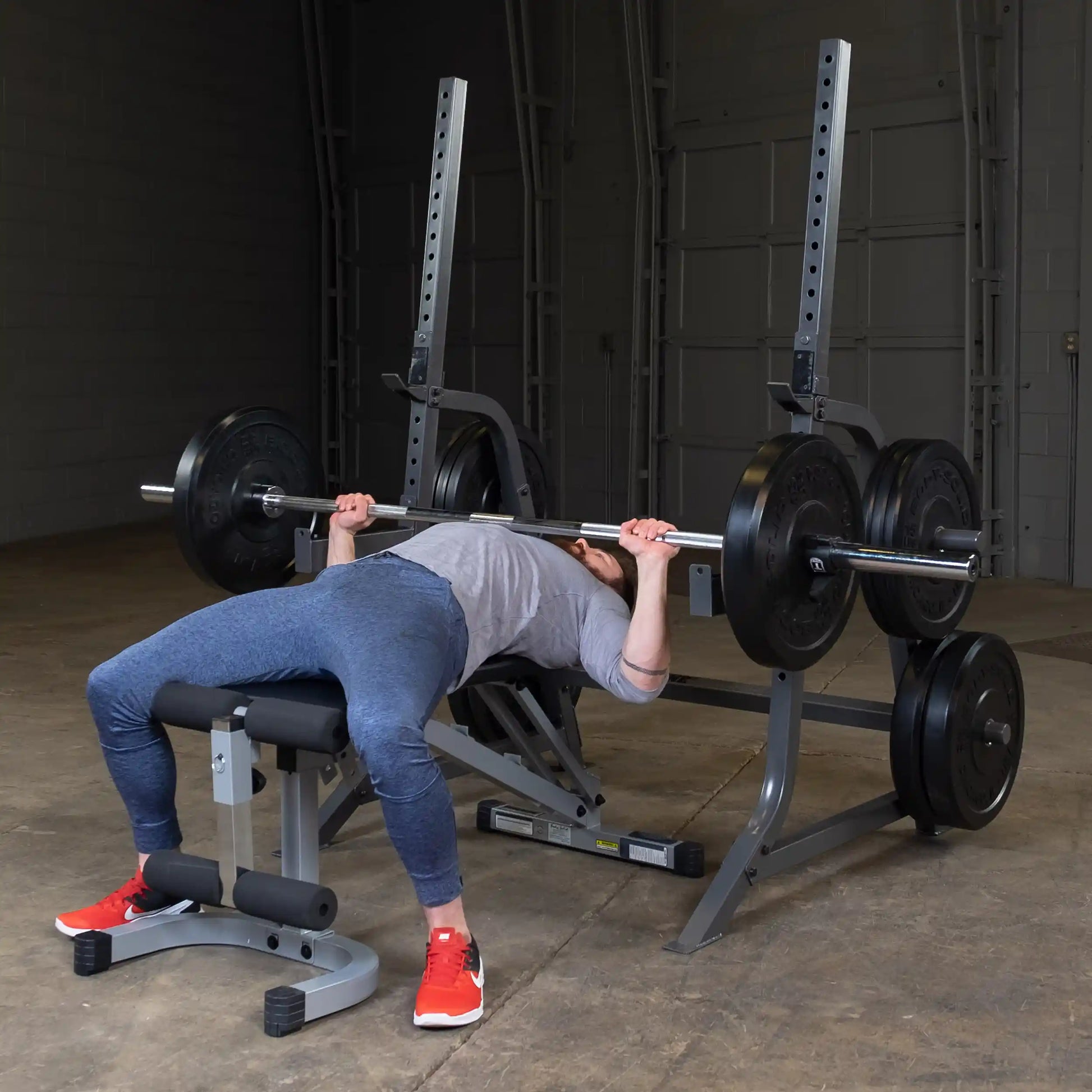 A person performing a bench press exercise in a gym setting, lying on a bench and lifting a barbell loaded with weight plates, wearing athletic clothing and red sneakers.