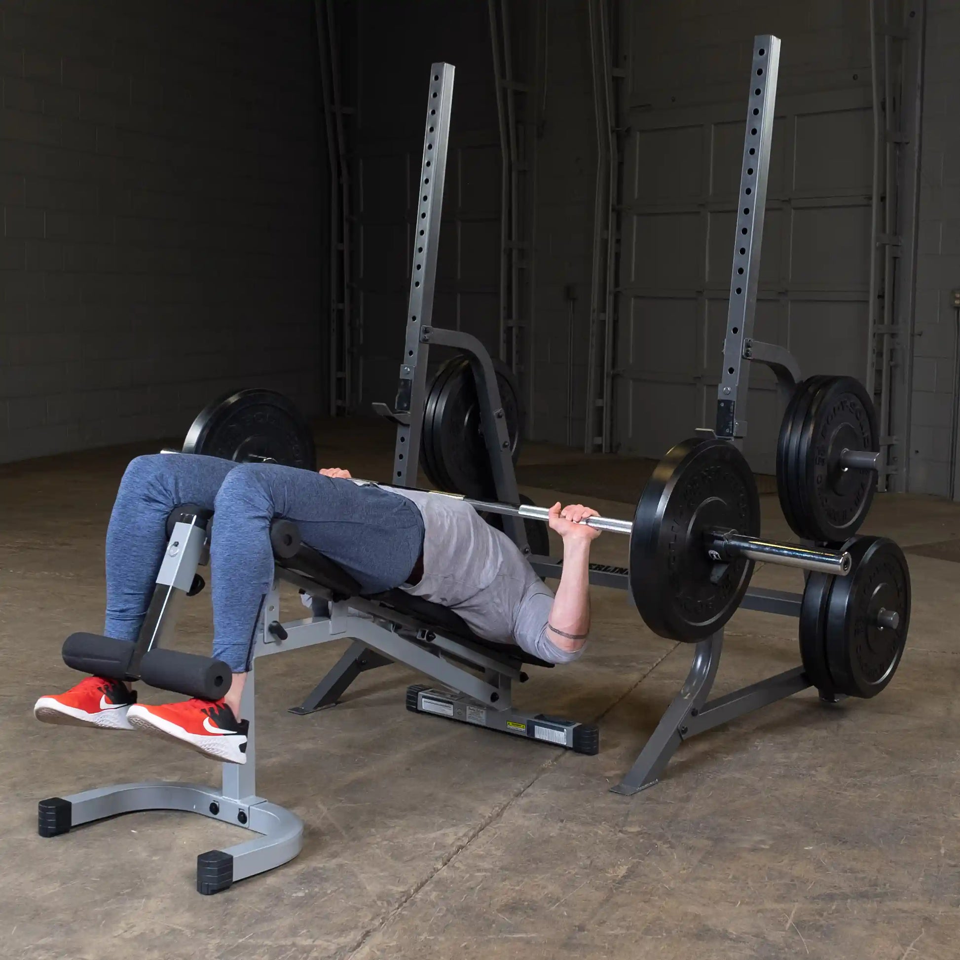Individual performing a bench press exercise with a barbell and weights in a gym setting, lying on a bench with feet supported.