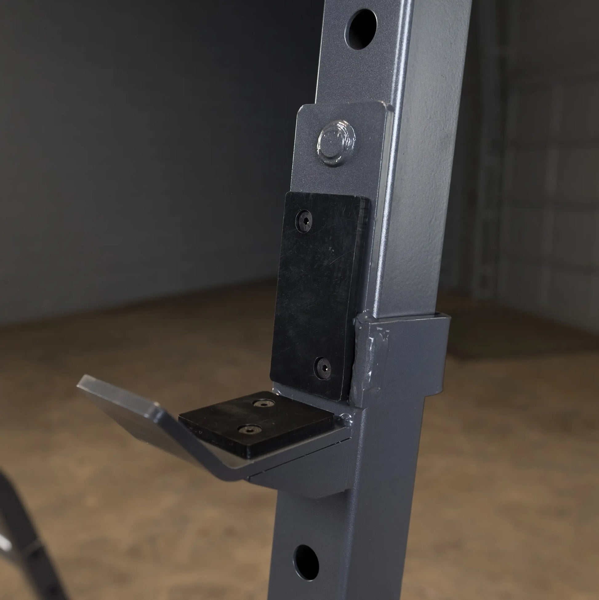 Close-up view of a gray metal gym rack with a black attachment plate and a horizontal support bracket, set against a blurred background.