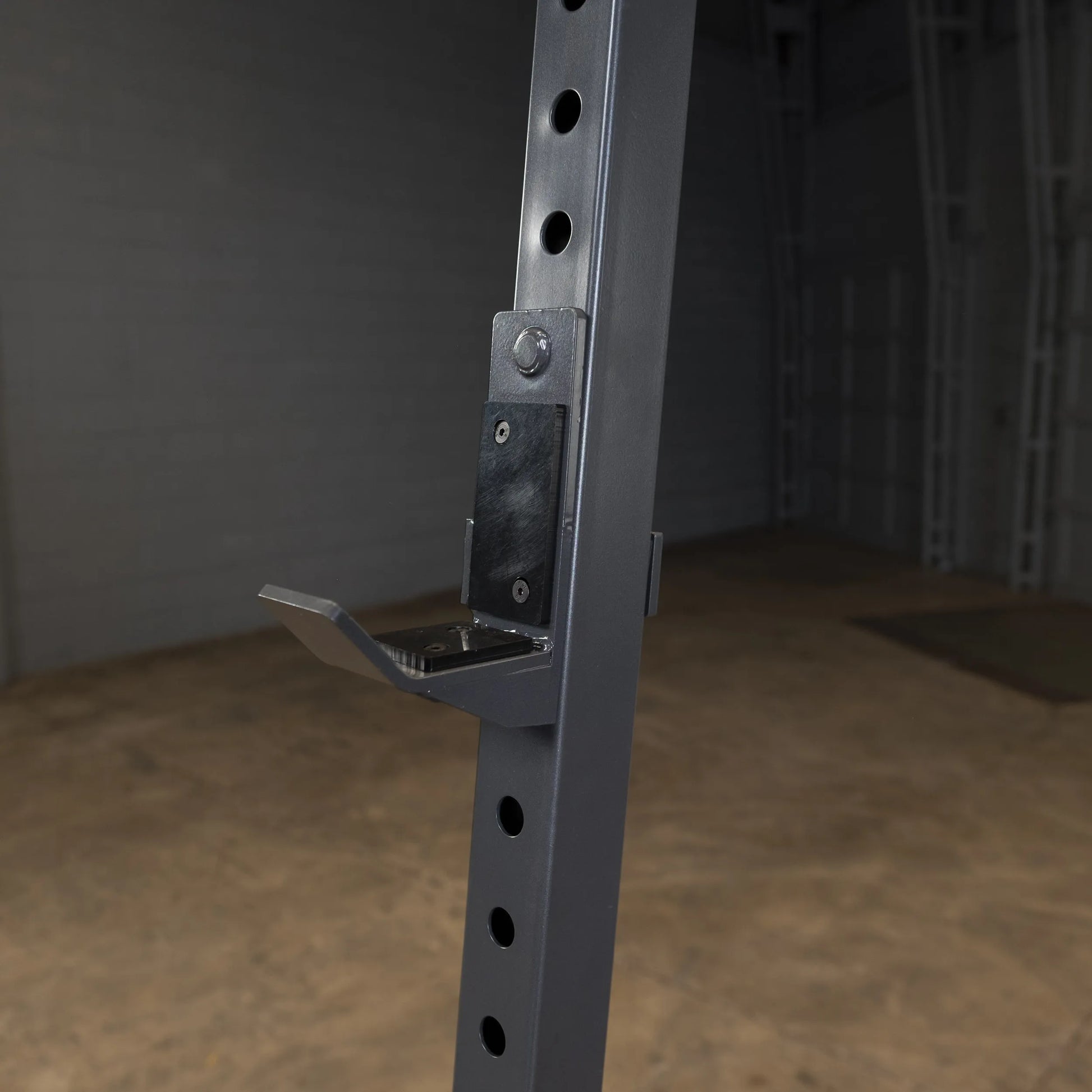 Close-up view of a metal weightlifting rack with adjustable hook and multiple holes for height adjustment, set against a blurred industrial background.