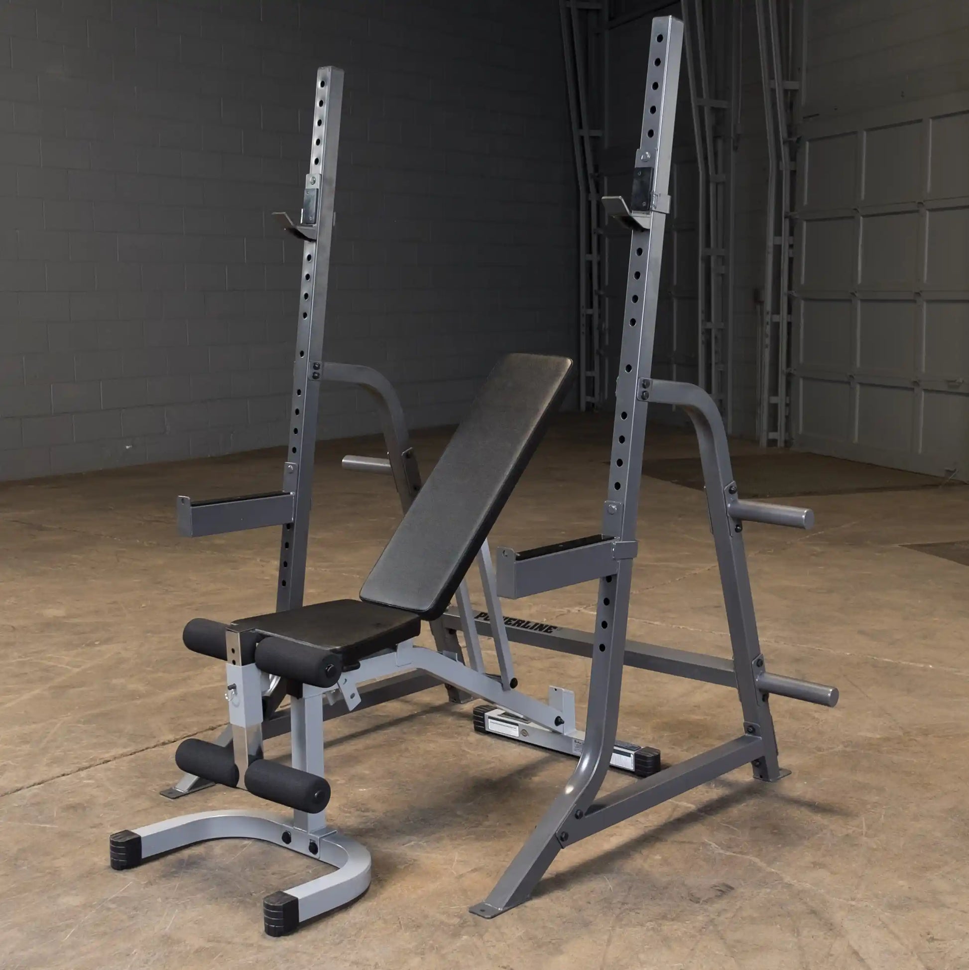 A gray weight bench with adjustable height and a barbell rack, situated on a concrete floor in a gym-like setting.