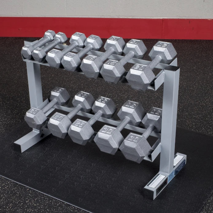 A rack of silver hexagonal dumbbells arranged neatly in two rows on a black rubber gym floor, with a red wall in the background.