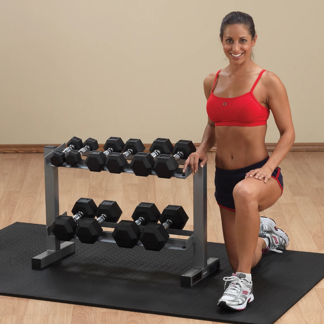 A fit woman in a red sports bra and black shorts kneels beside a rack of dumbbells on a gym mat, smiling at the camera.