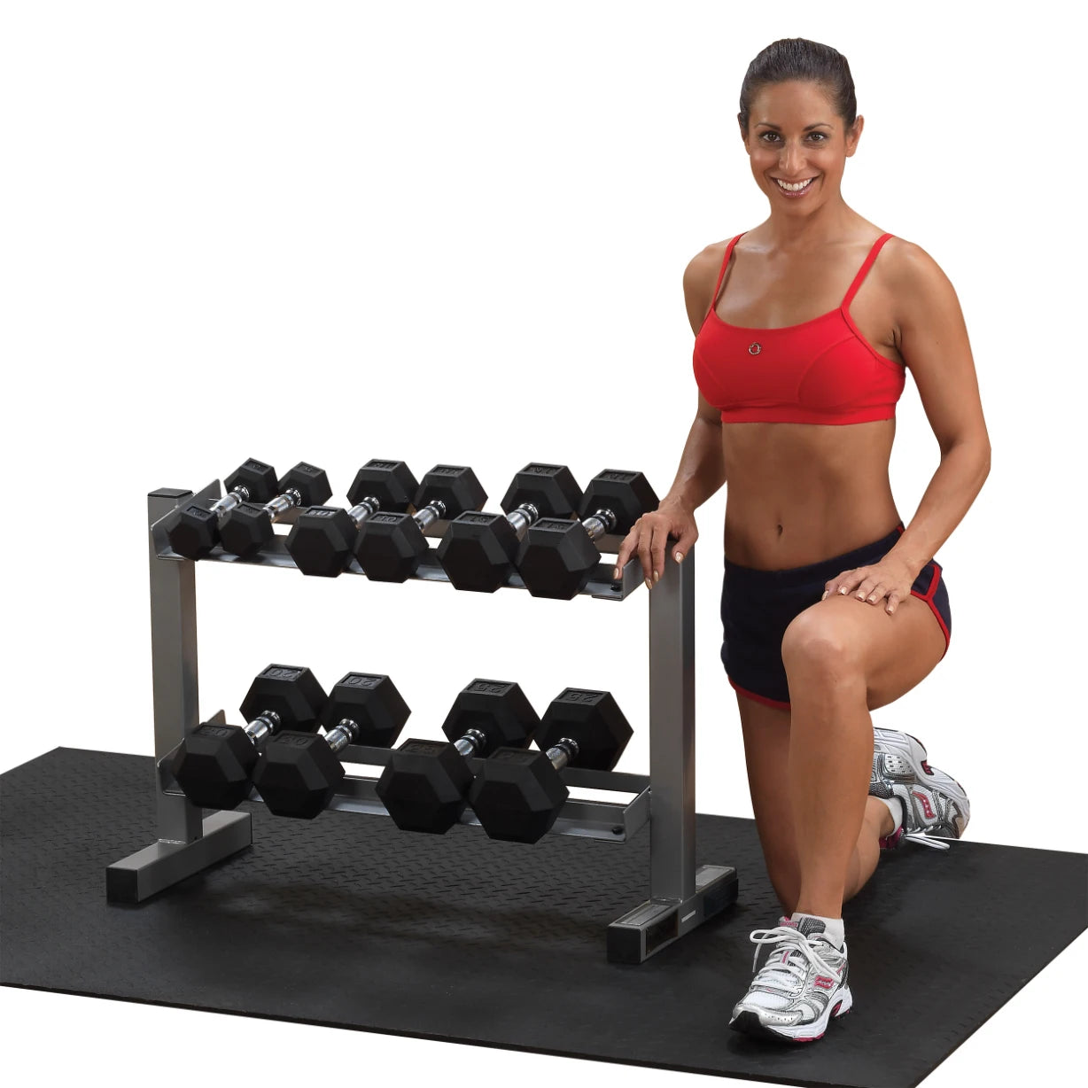 A fit woman in a red sports bra and black shorts kneels beside a rack of hexagonal dumbbells on a rubber mat, smiling confidently.