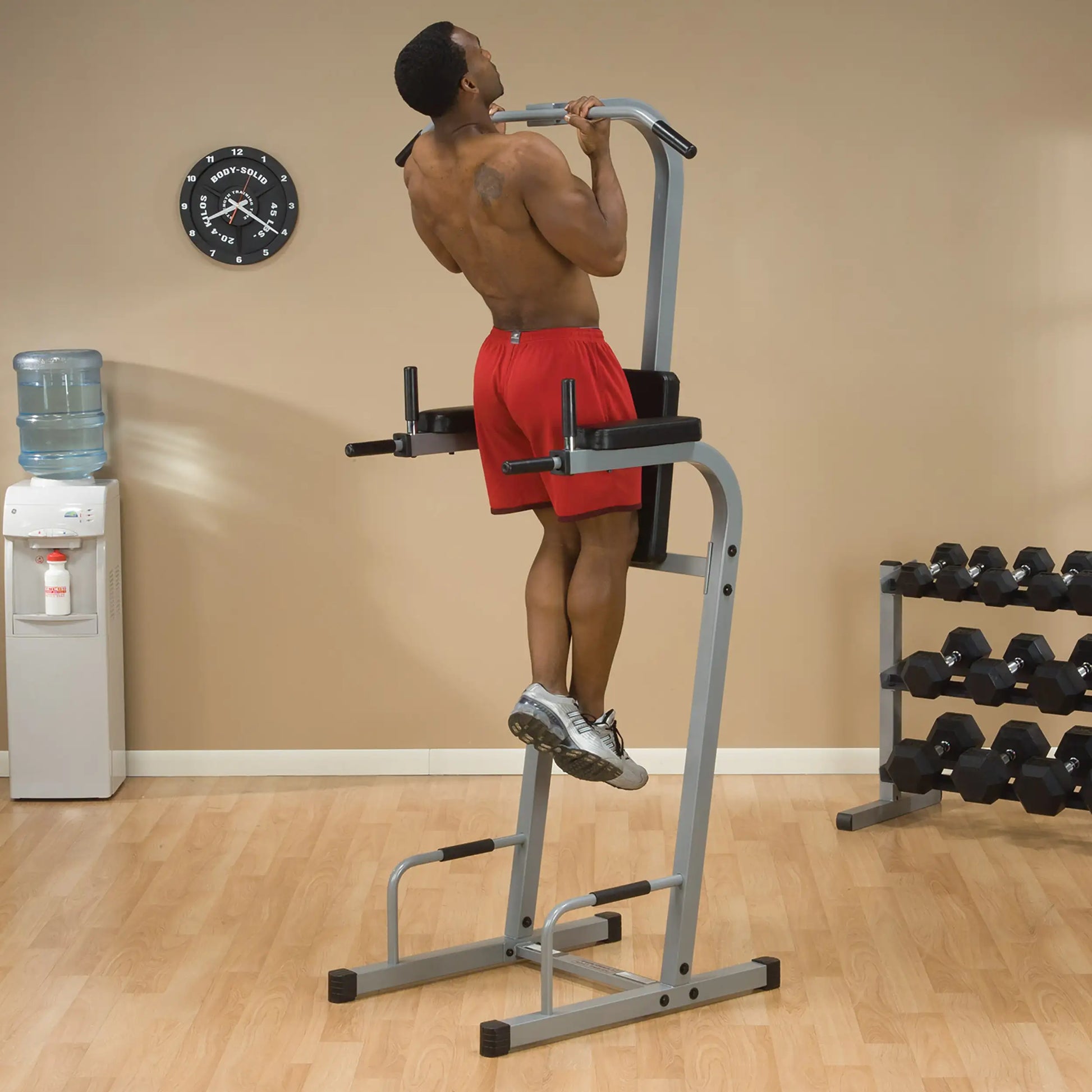 A person performing a pull-up on a vertical bar in a gym setting, wearing red shorts. In the background, there is a water cooler, a clock on the wall, and a rack of dumbbells.