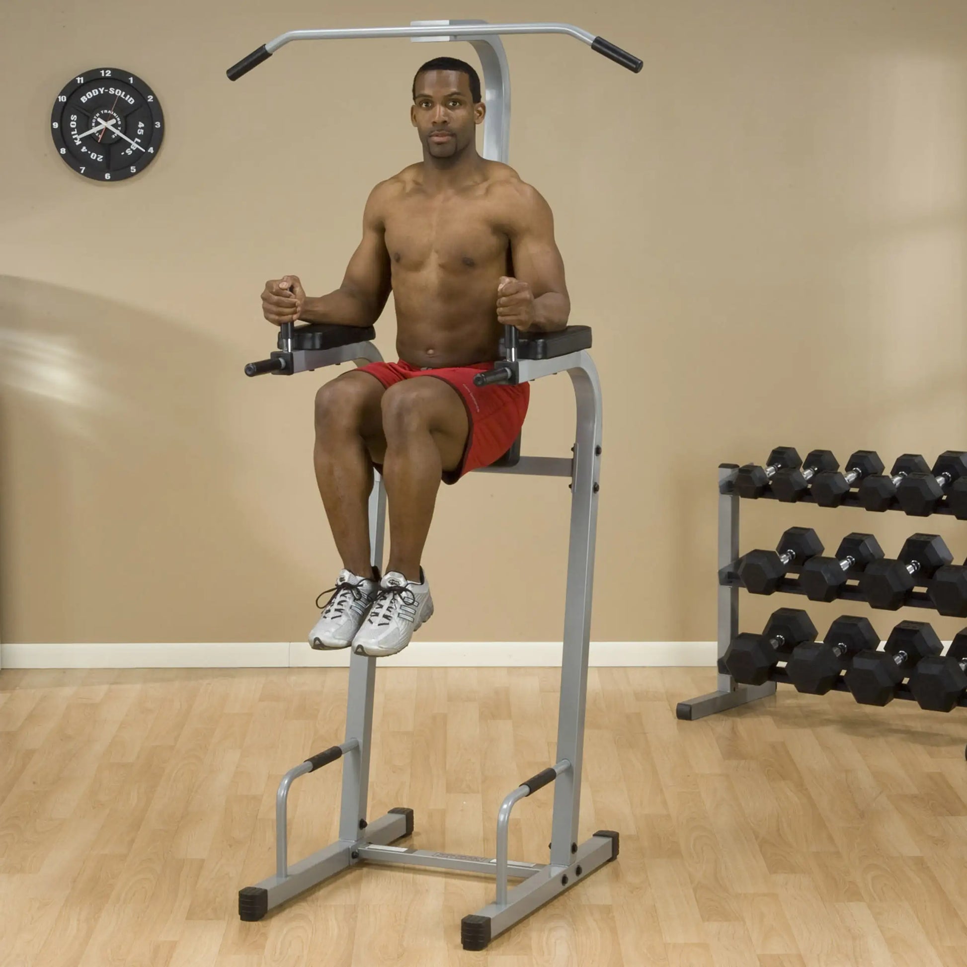 A shirtless man performing a dip exercise on a vertical gym apparatus in a fitness studio, with a clock on the wall and a rack of dumbbells in the background.