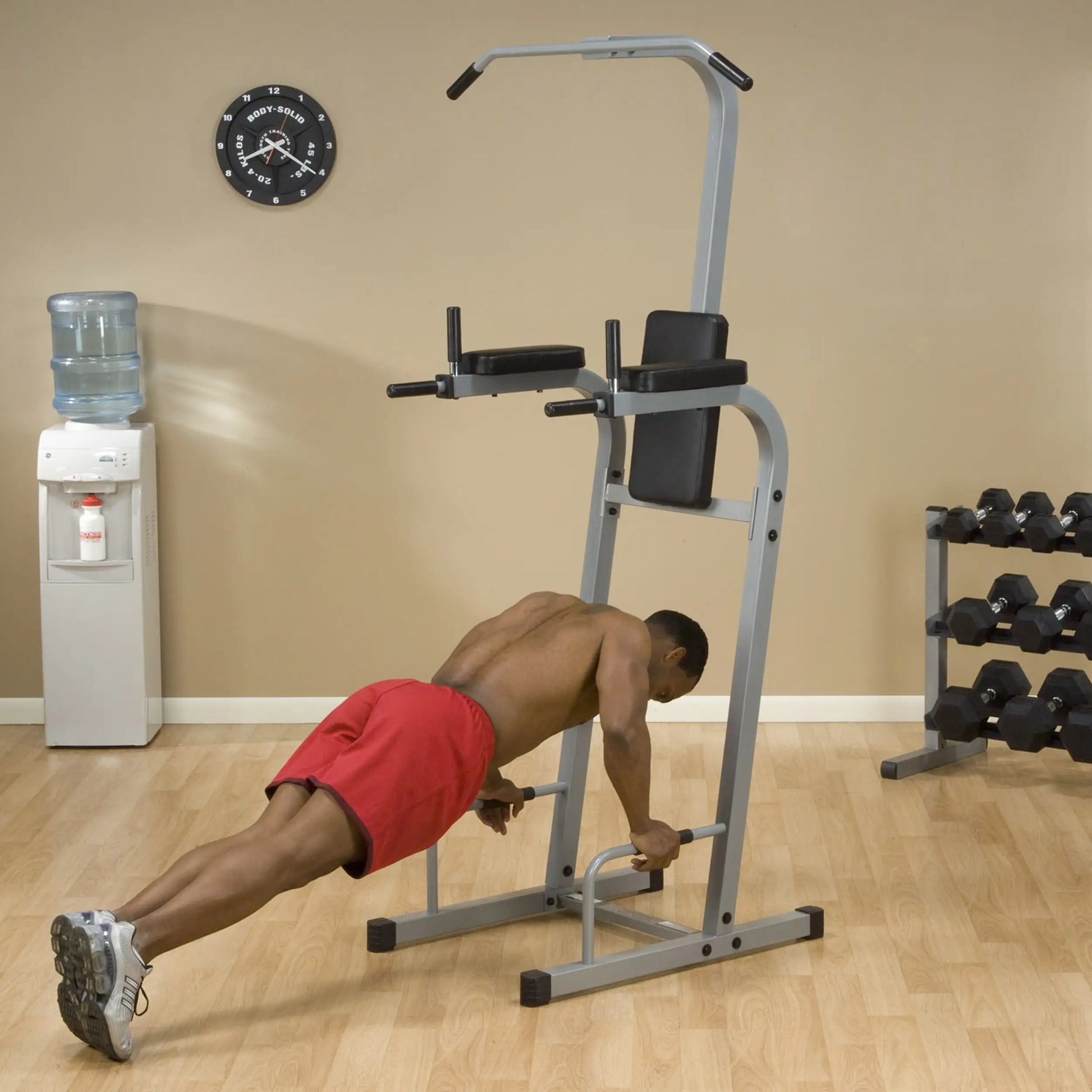 A person performing push-ups on a fitness apparatus in a gym setting, with a water cooler and a rack of dumbbells in the background.