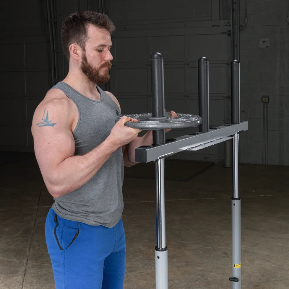 A man with a beard wearing a gray tank top and blue pants is preparing to place a weight plate on a barbell rack in a gym setting.