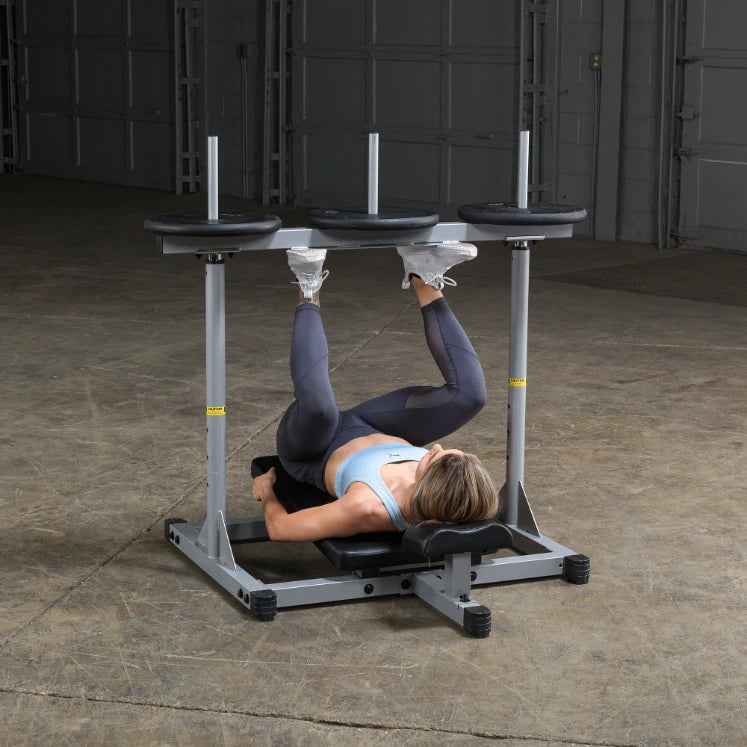 A person performing an exercise on a specialized fitness machine, lying on their back with legs raised and supported by padded bars, in a spacious gym environment.