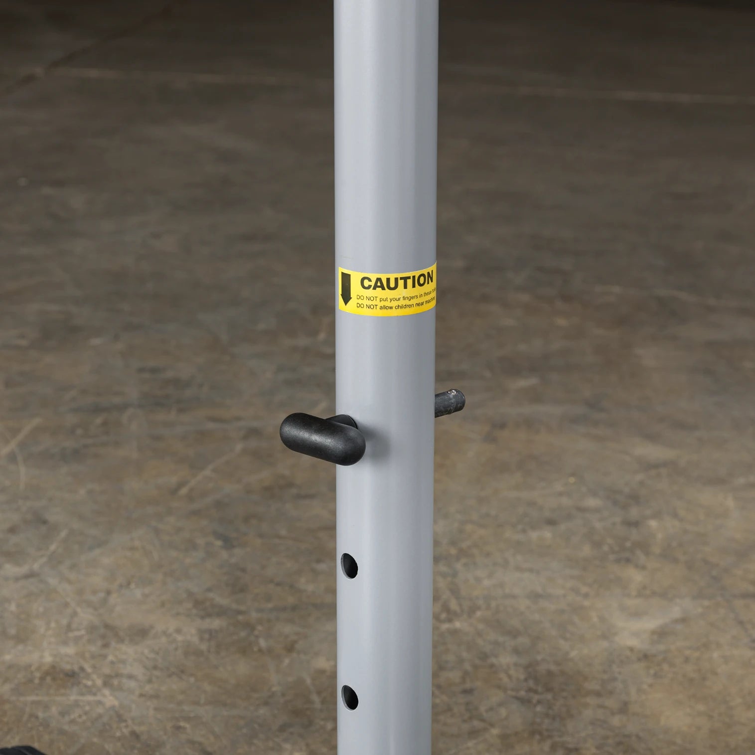 A close-up of a gray metal pole with a caution label reading CAUTION: DO NOT put your fingers in holes. DO NOT allow children near attached to it. The pole features a black handle and several holes along its length, set against a concrete floor.