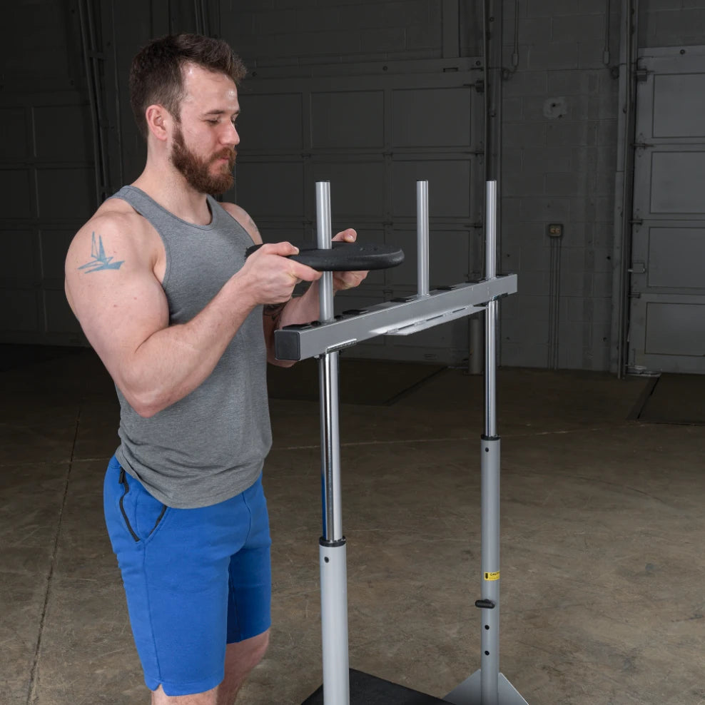 A man with a beard and tattoos is preparing to place a weight plate on a barbell rack in a gym setting. He is wearing a gray tank top and blue shorts, standing on a concrete floor.