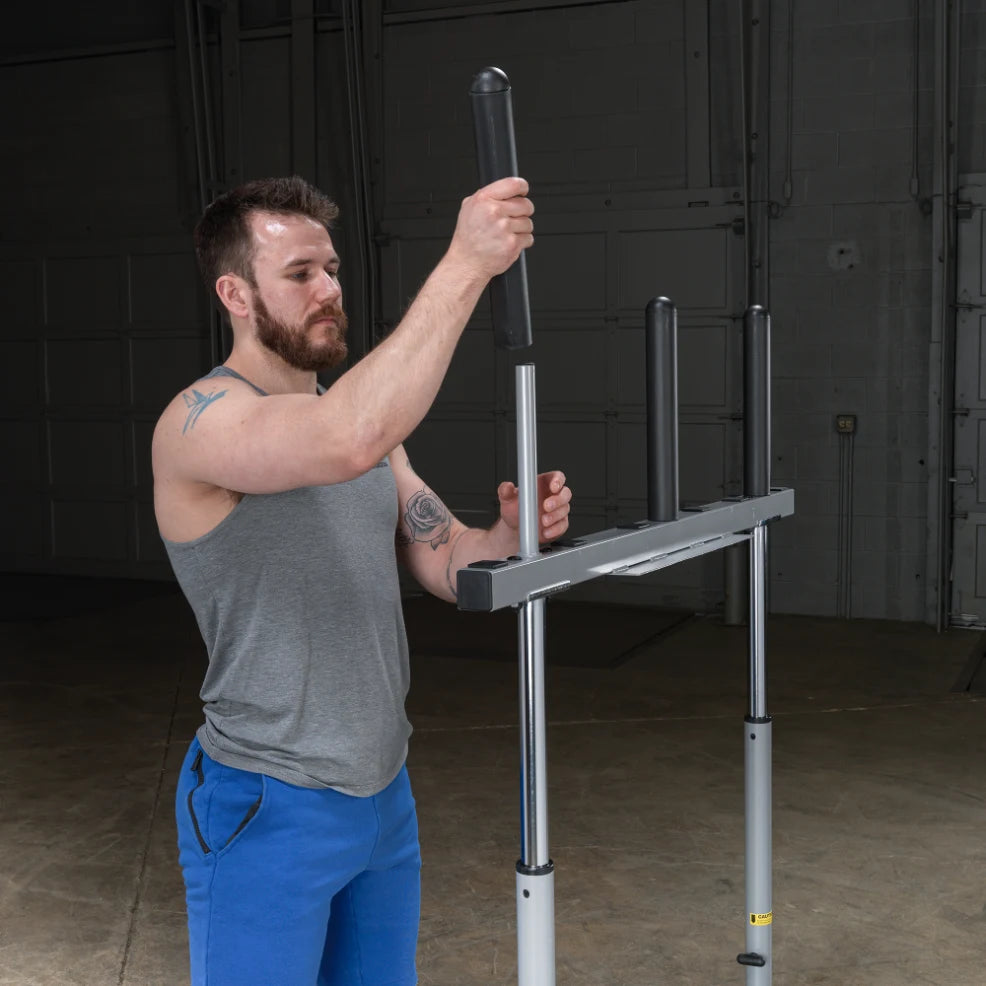 A man in a gray tank top and blue shorts adjusts the height of a pull-up bar in a gym setting.