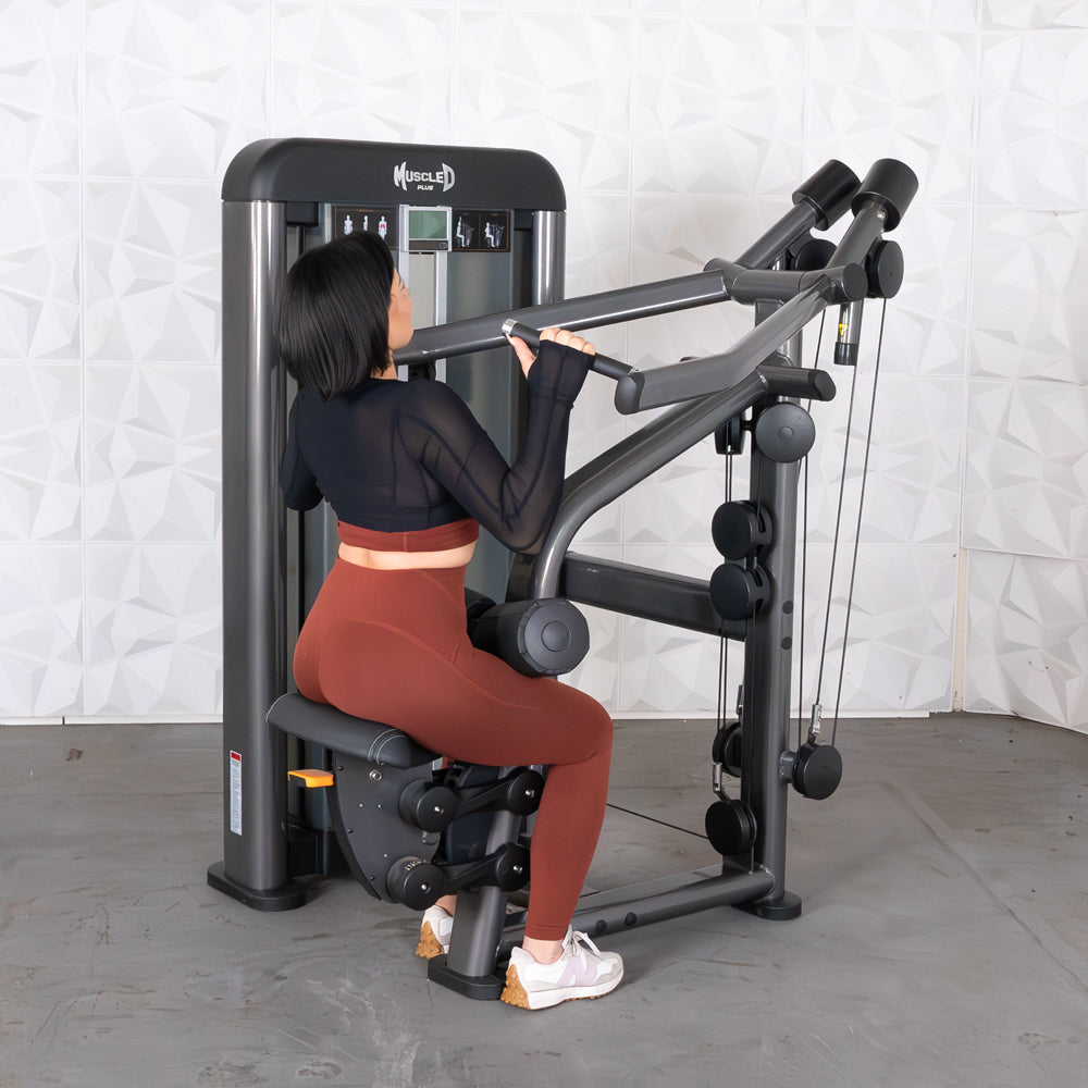 Woman using MuscleD lat pulldown machine in gym, wearing black top and red leggings.