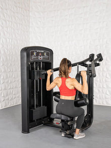 Woman using Muscle D lat pulldown machine in gym, wearing red top and black leggings.