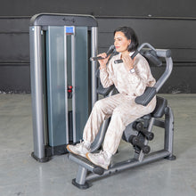 Woman using Muscle D shoulder press machine in a beige tracksuit.