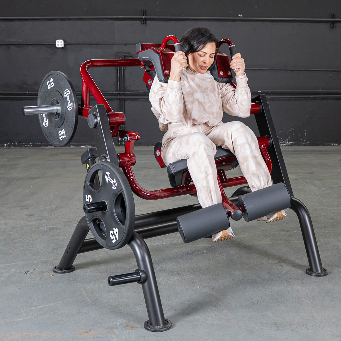 Woman in beige workout outfit using a red weight machine for leg exercises in a gym.