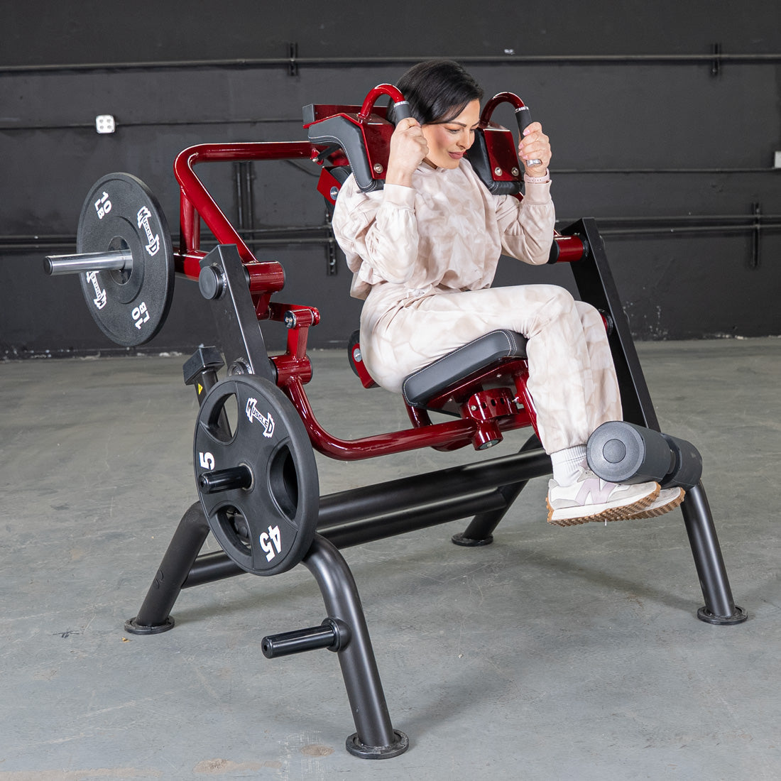 Woman using red plate-loaded shoulder press machine in a gym, wearing beige workout set.