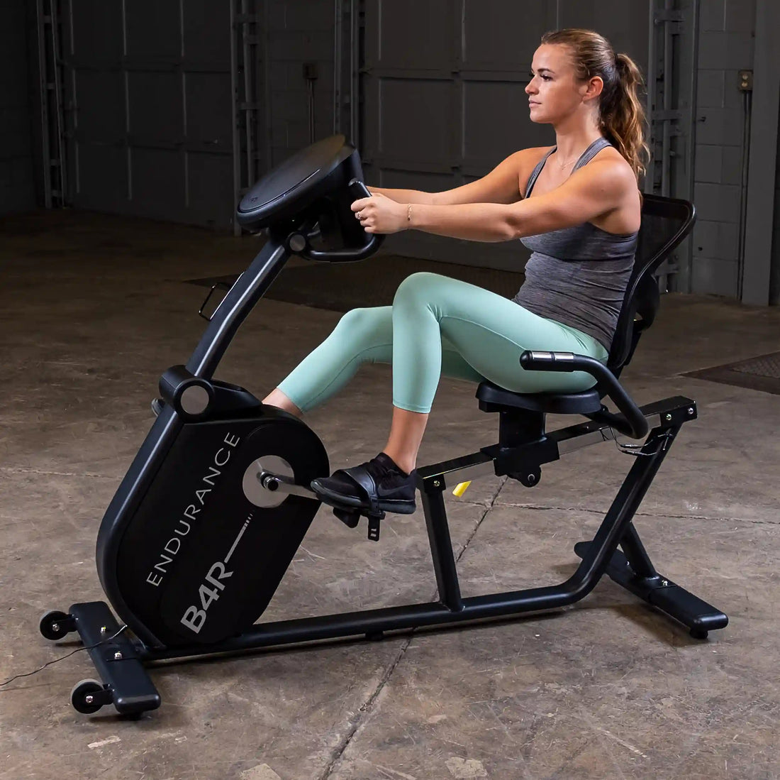 Woman exercising on a Body-Solid Endurance B4RB recumbent bike in an industrial gym space, seen from the side