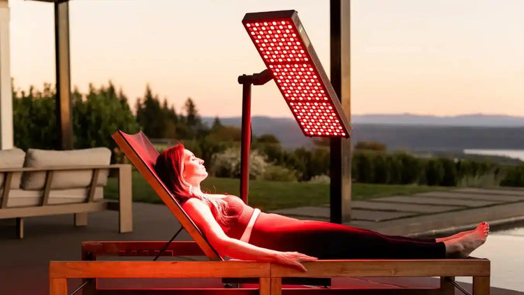 Person lying on a wooden lounge chair under a red light therapy panel with a scenic background.