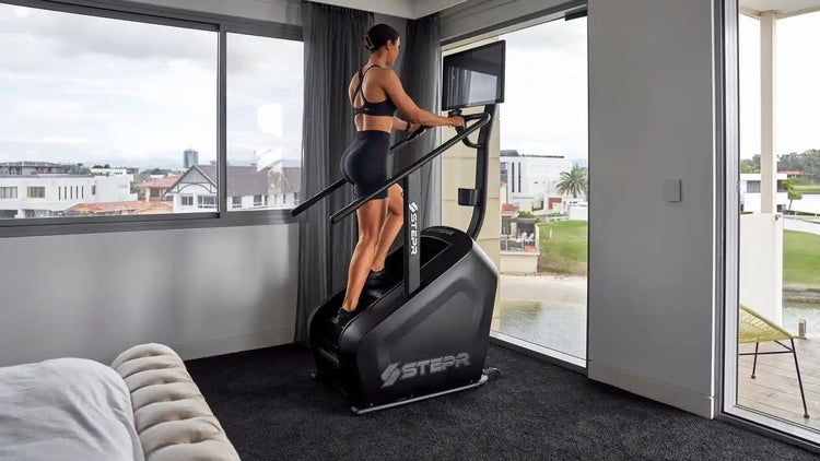 Woman on STEPR stair climber cardio machine in a home setting surrounded by glass windows