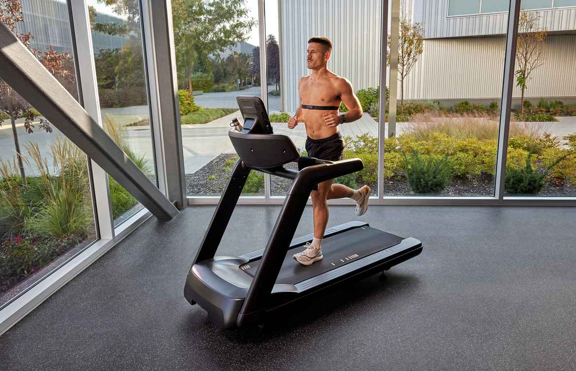 Man running on a black treadmill with digital display in a modern gym with floor-to-ceiling windows overlooking landscaped grounds.
