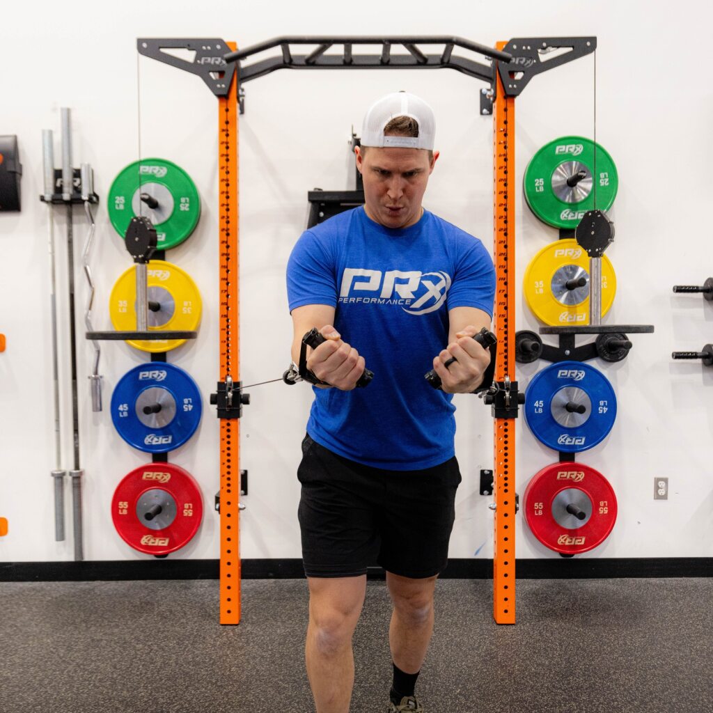 Man in blue PRx shirt performing dumbbell exercise in gym with weight plates