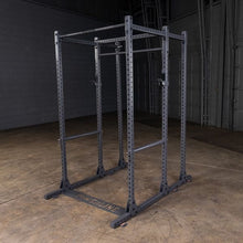 A black power rack with multiple adjustable bars, positioned in a spacious gym environment with a concrete floor and gray walls.