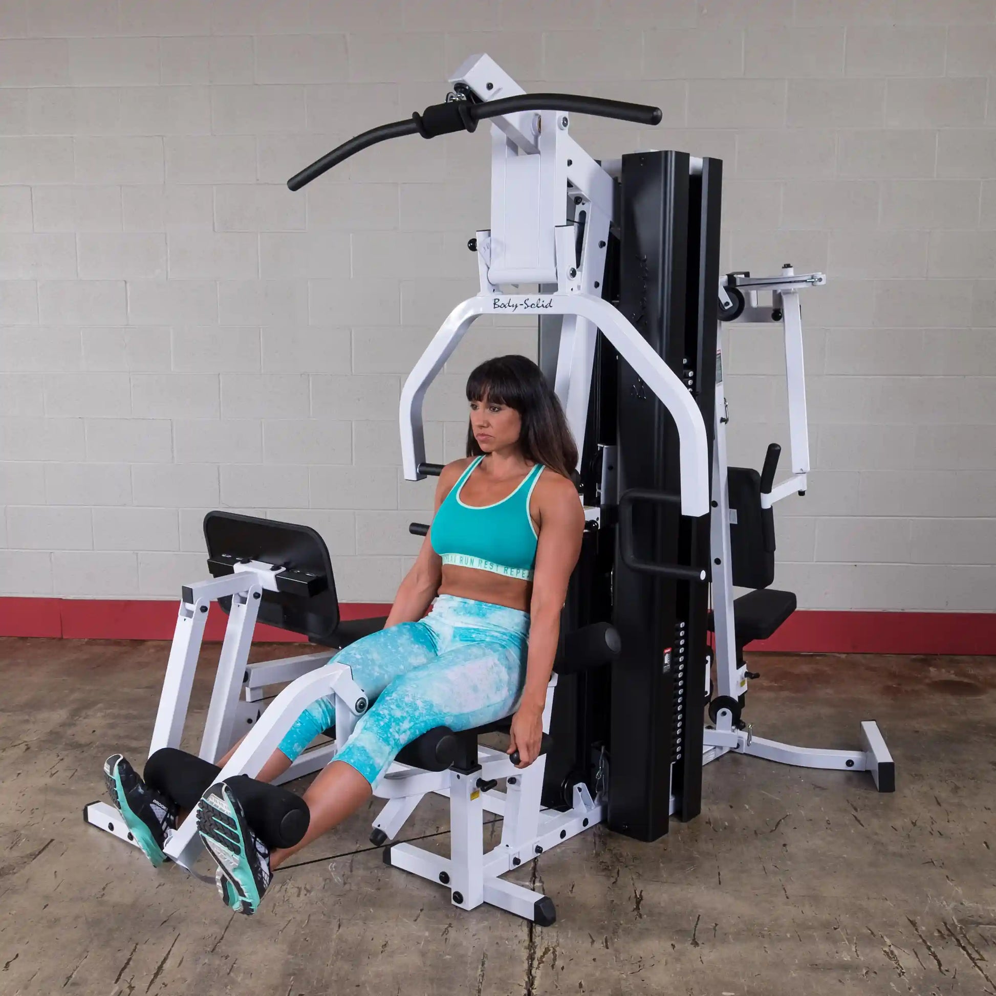 Woman using Body-Solid gym machine in teal workout outfit.