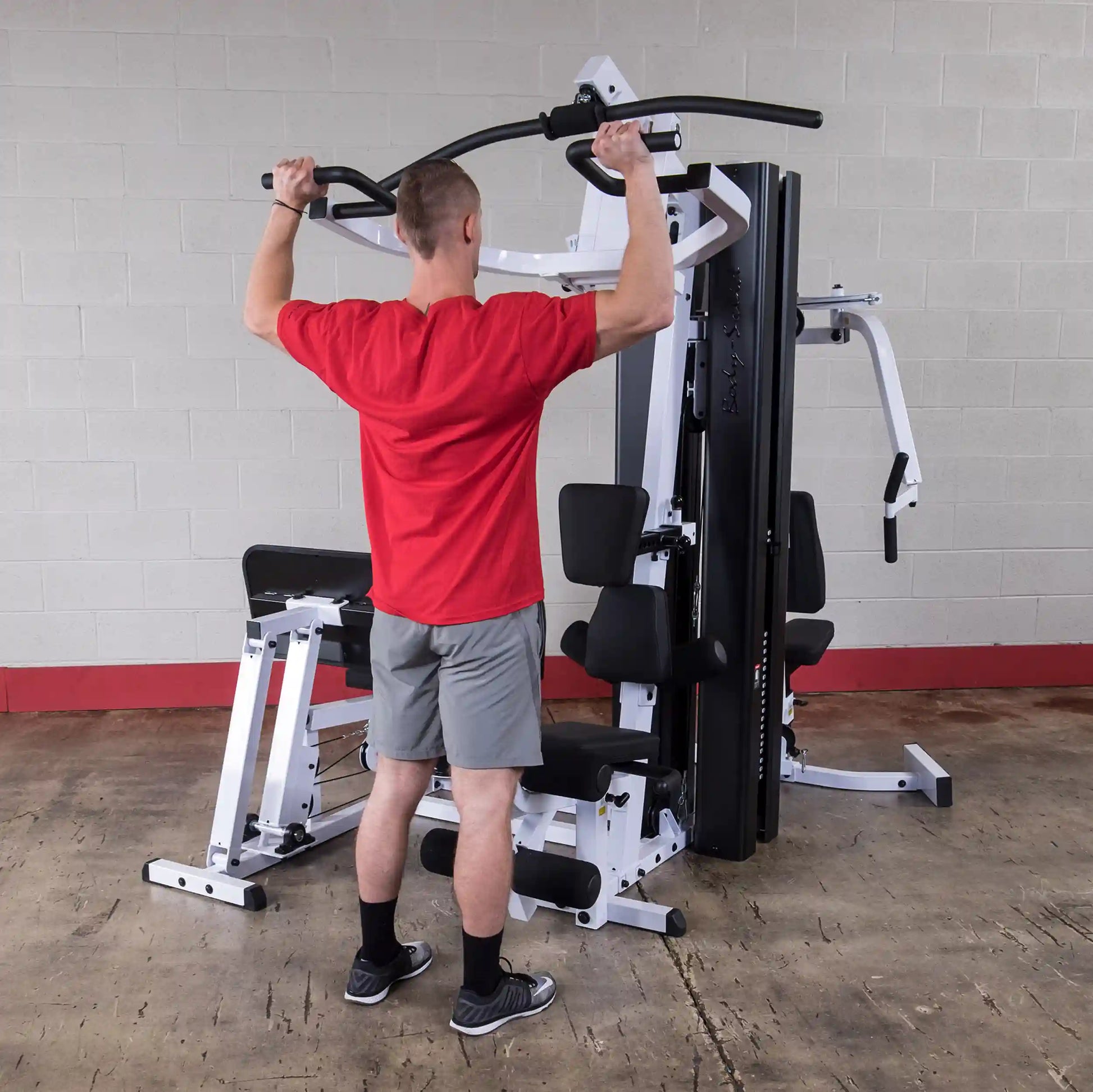 Man using home gym machine in red shirt and gray shorts for upper body workout.