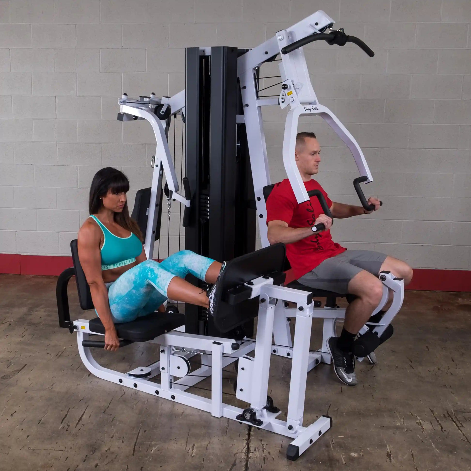 Two people using a Body-Solid home gym, one doing leg presses, the other performing chest presses.