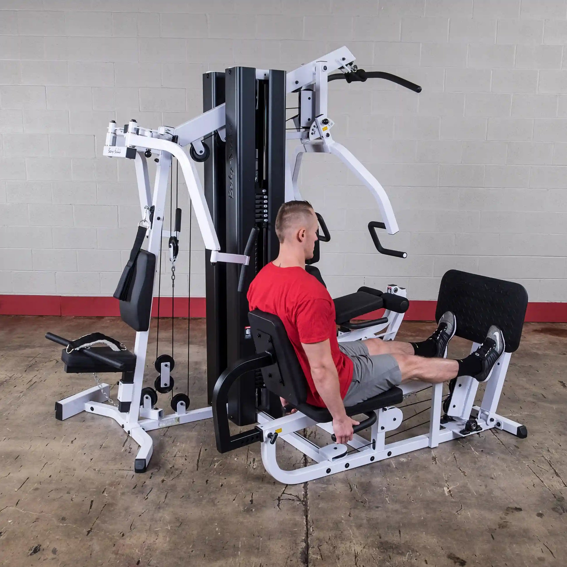 Man in red shirt using a Body-Solid multi-station gym machine for leg presses.