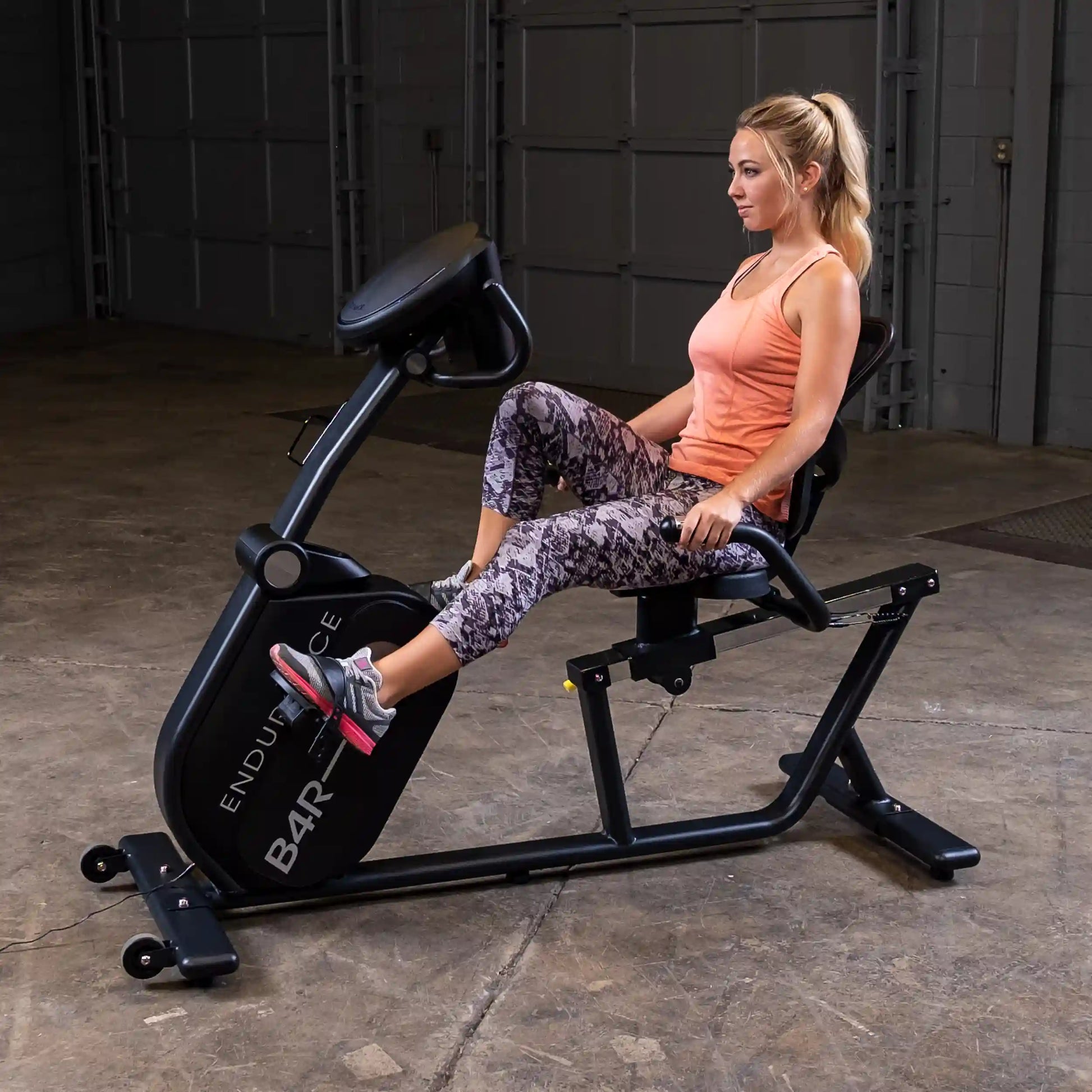 Woman using Endurance B4R recumbent exercise bike in industrial gym setting.
