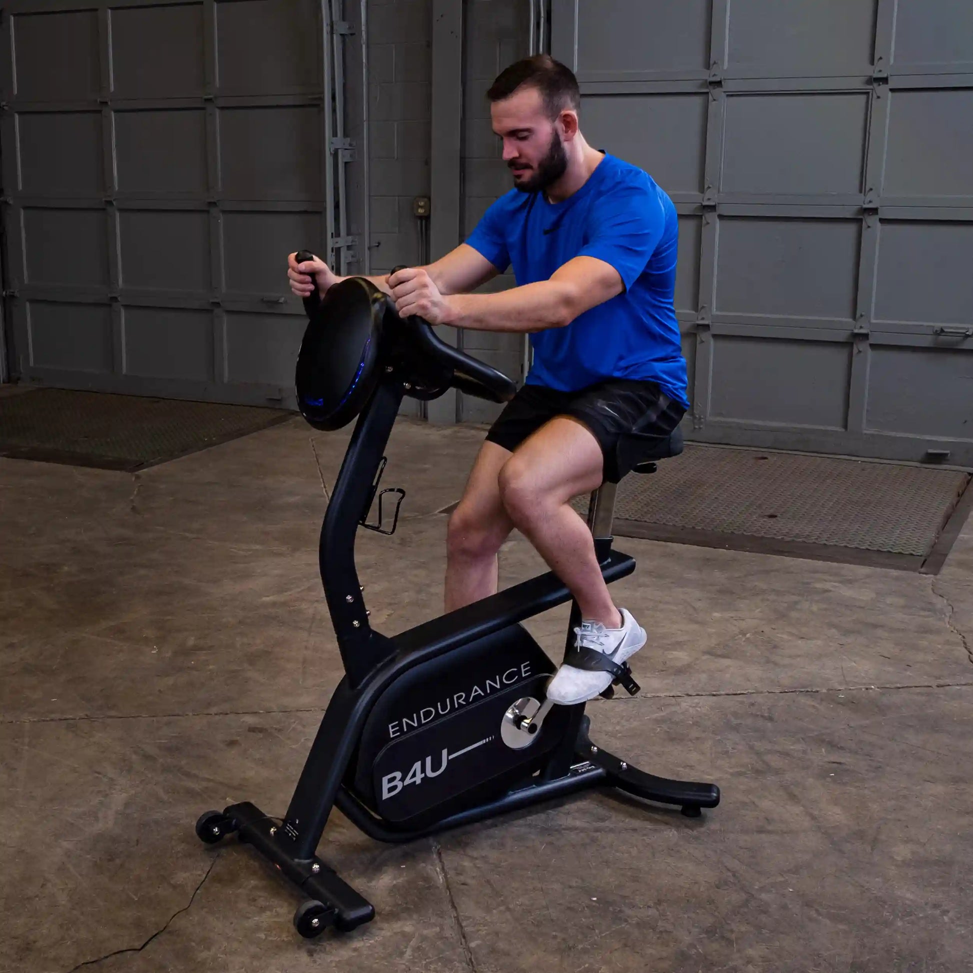 Man exercising on Endurance B4U stationary bike in a garage, wearing a blue shirt and black shorts.