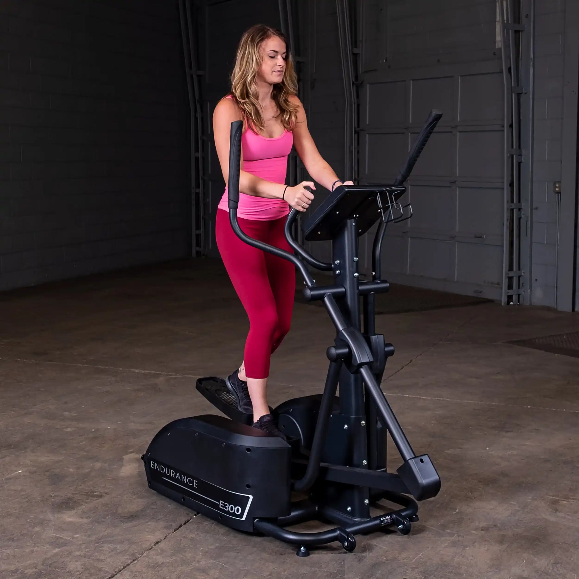 Woman using Endurance E300 elliptical in a gym, wearing pink and red workout clothes.