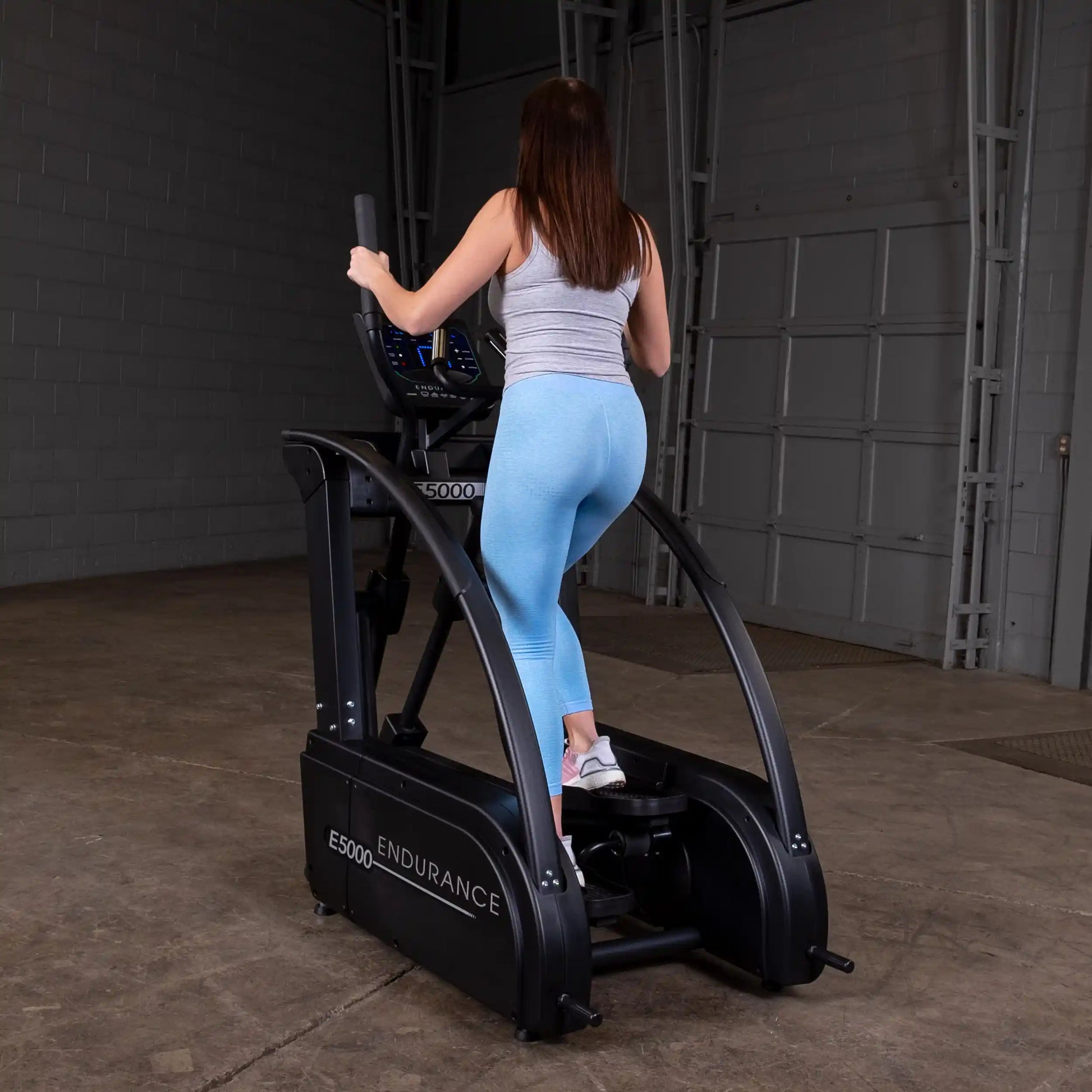 Woman exercising on Endurance E5000 elliptical, wearing blue leggings in a gym setting.