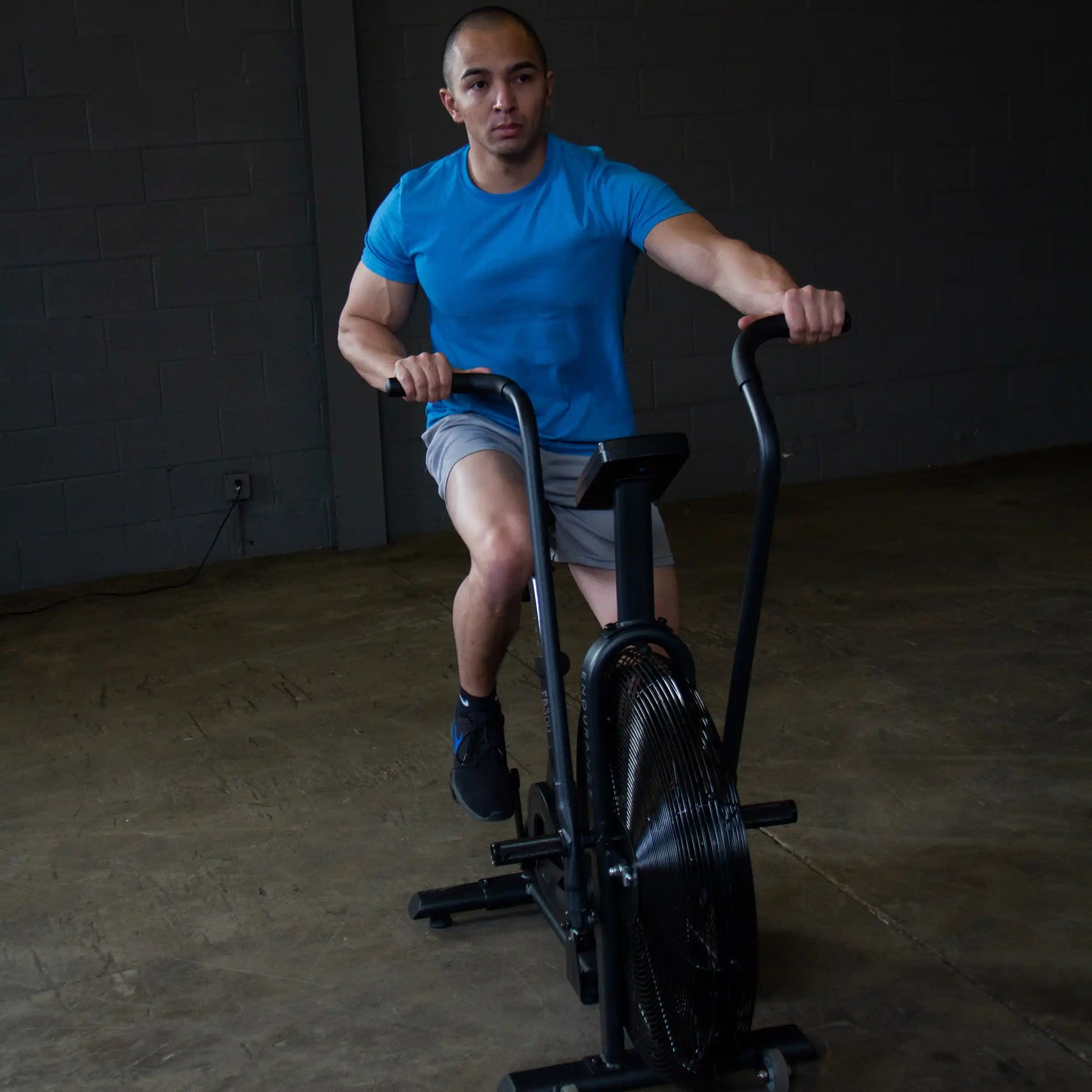 Man in blue shirt exercises on a black air bike in a dimly lit gym.