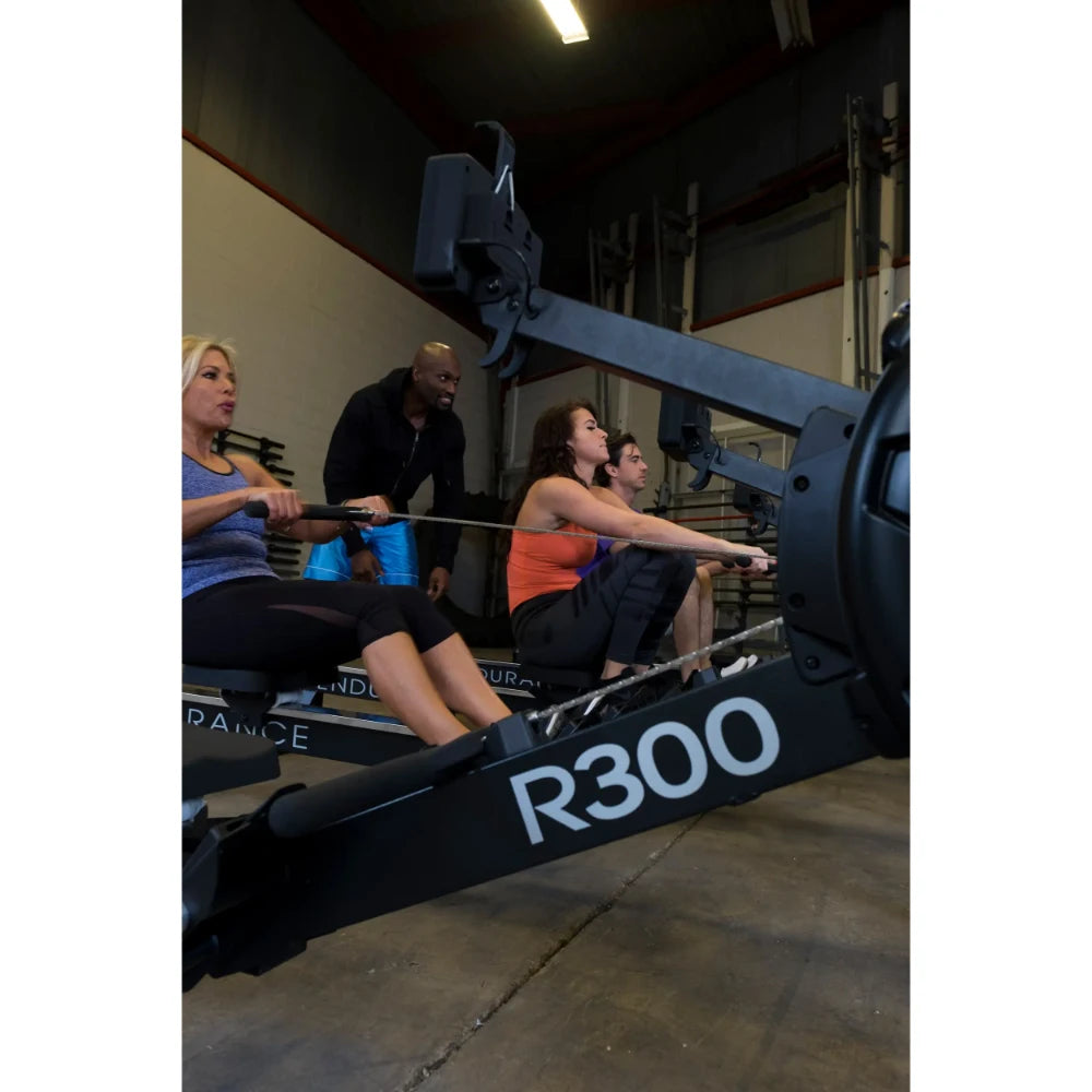 Group rowing on R300 machines in a gym, led by a trainer.