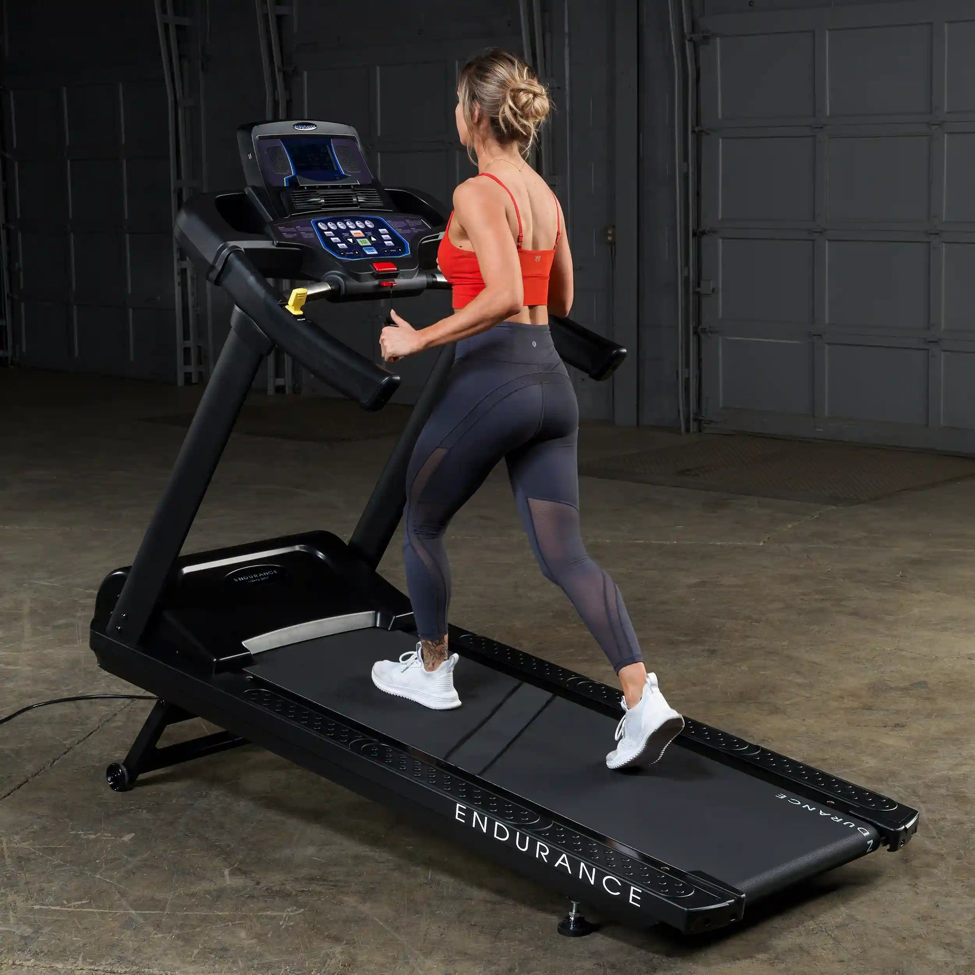Woman running on Endurance treadmill in a garage, wearing a red top and black leggings.