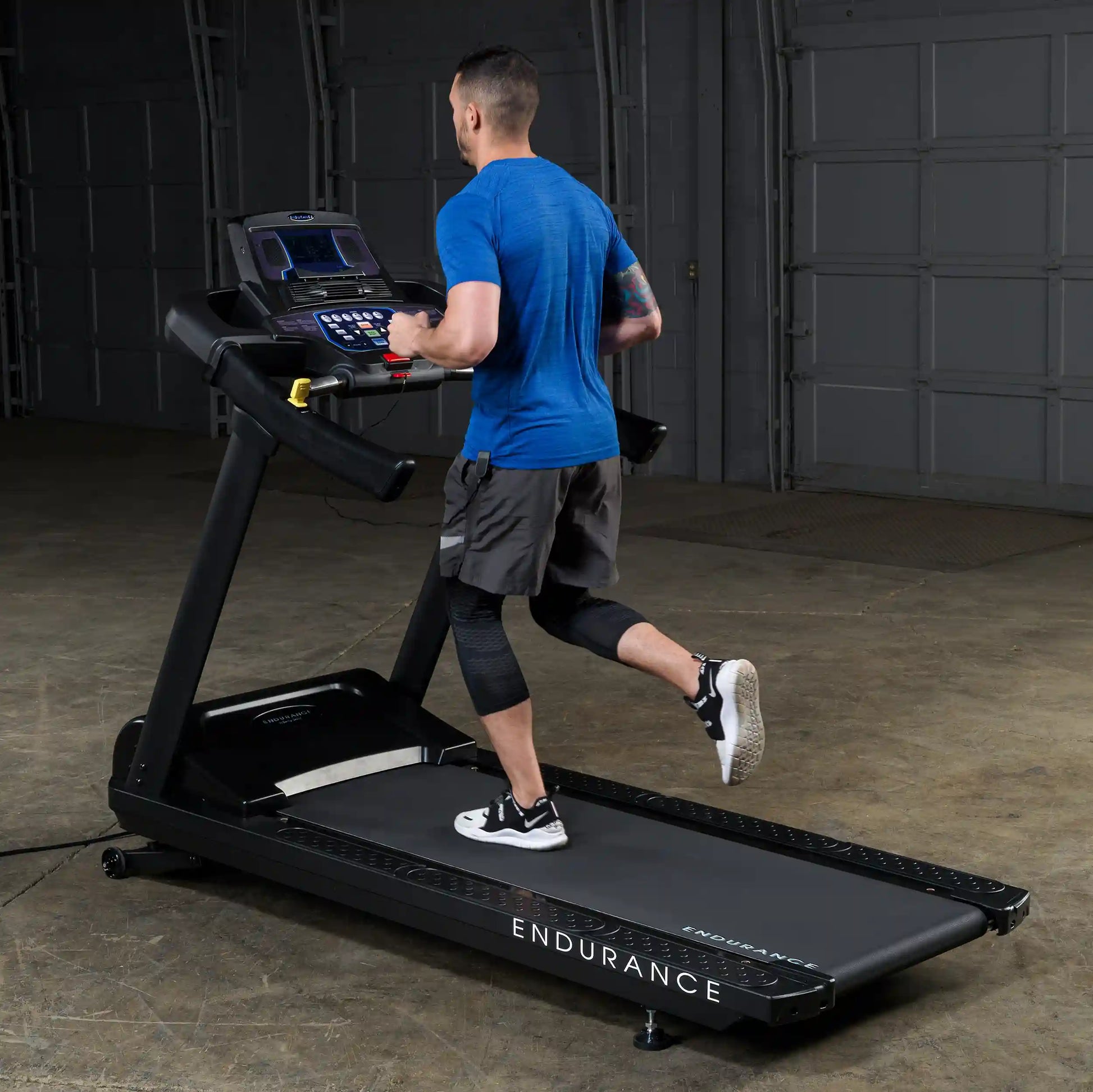 Man running on Endurance treadmill in a blue shirt, showcasing fitness equipment in a garage setting.