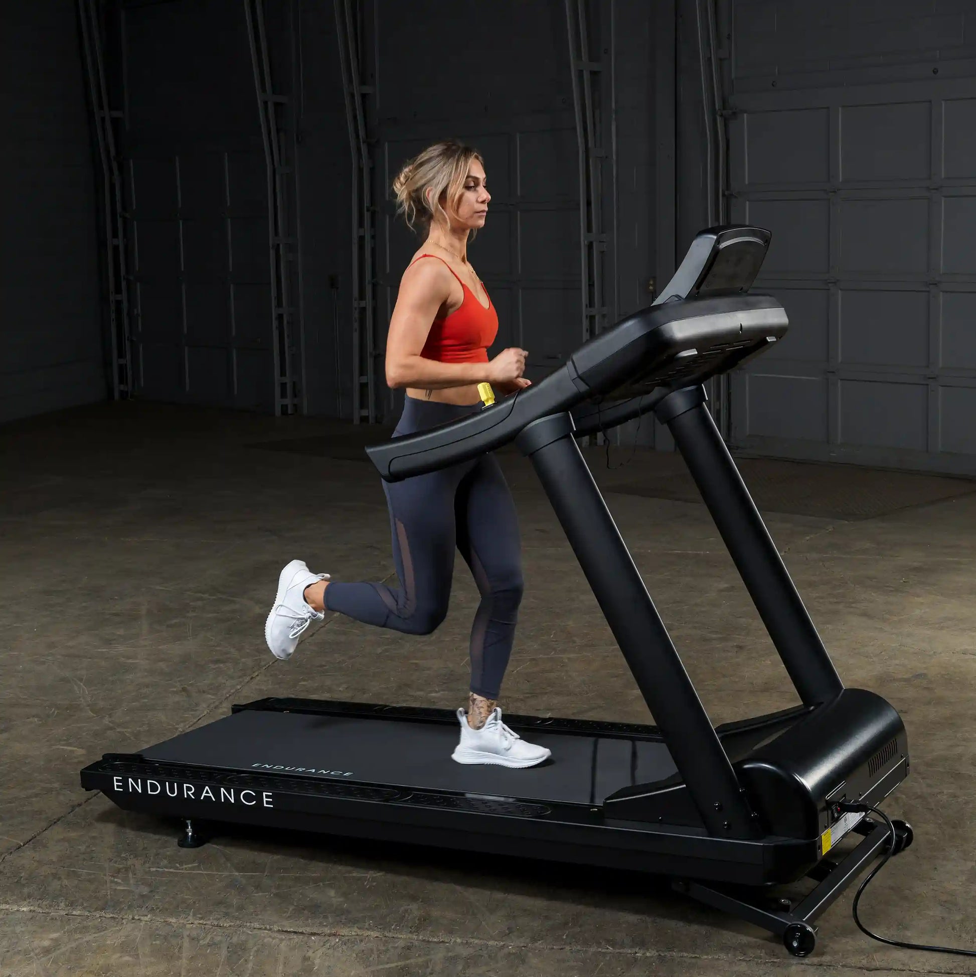 Woman running on black Endurance treadmill indoors, wearing red top and dark leggings.
