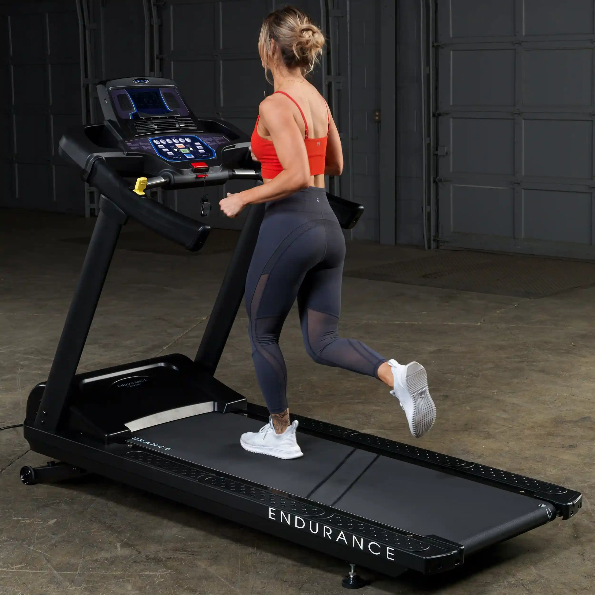 Woman running on Endurance treadmill in a gym, wearing red top and black leggings.