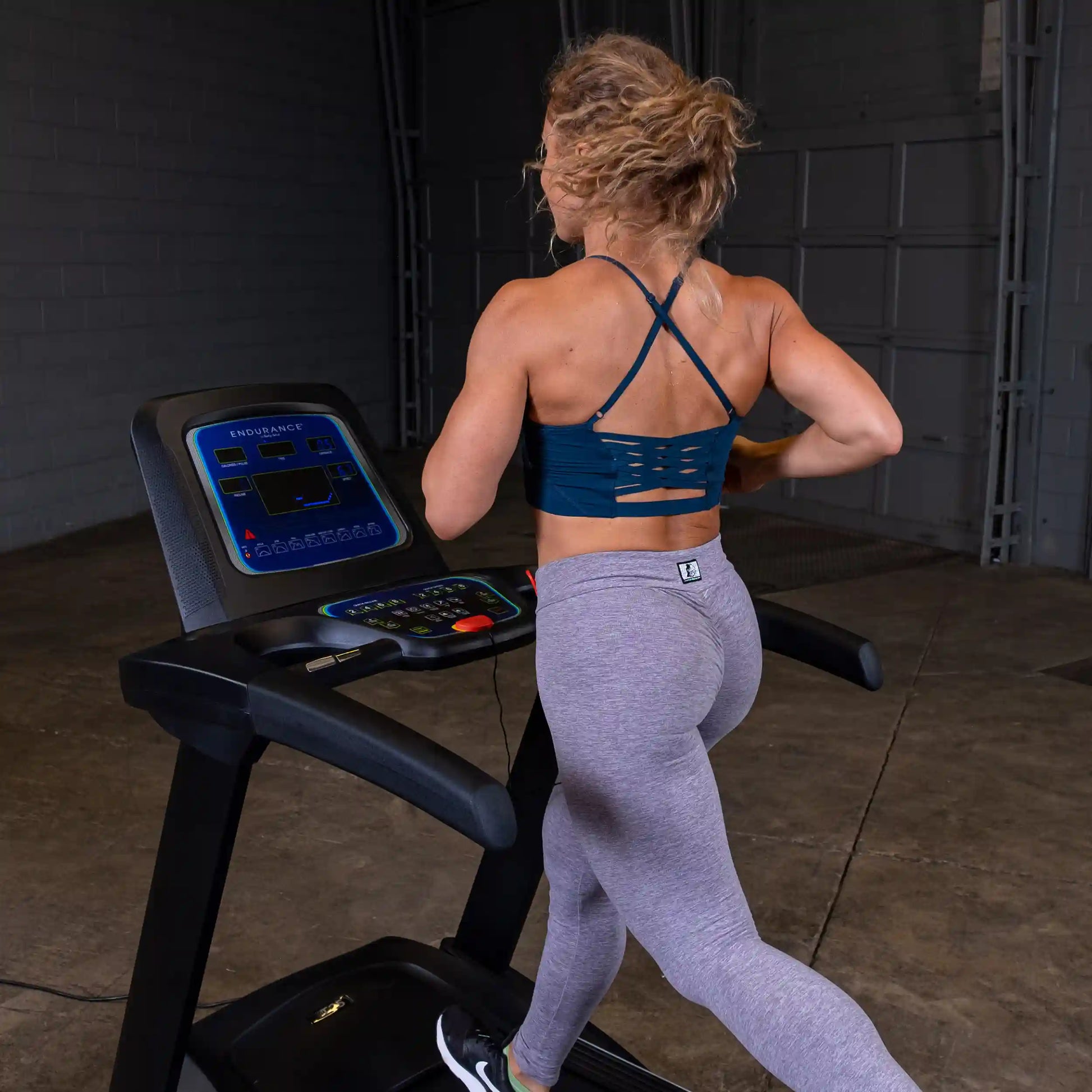 Woman running on a black Endurance treadmill in a gym wearing a blue sports bra and gray leggings.