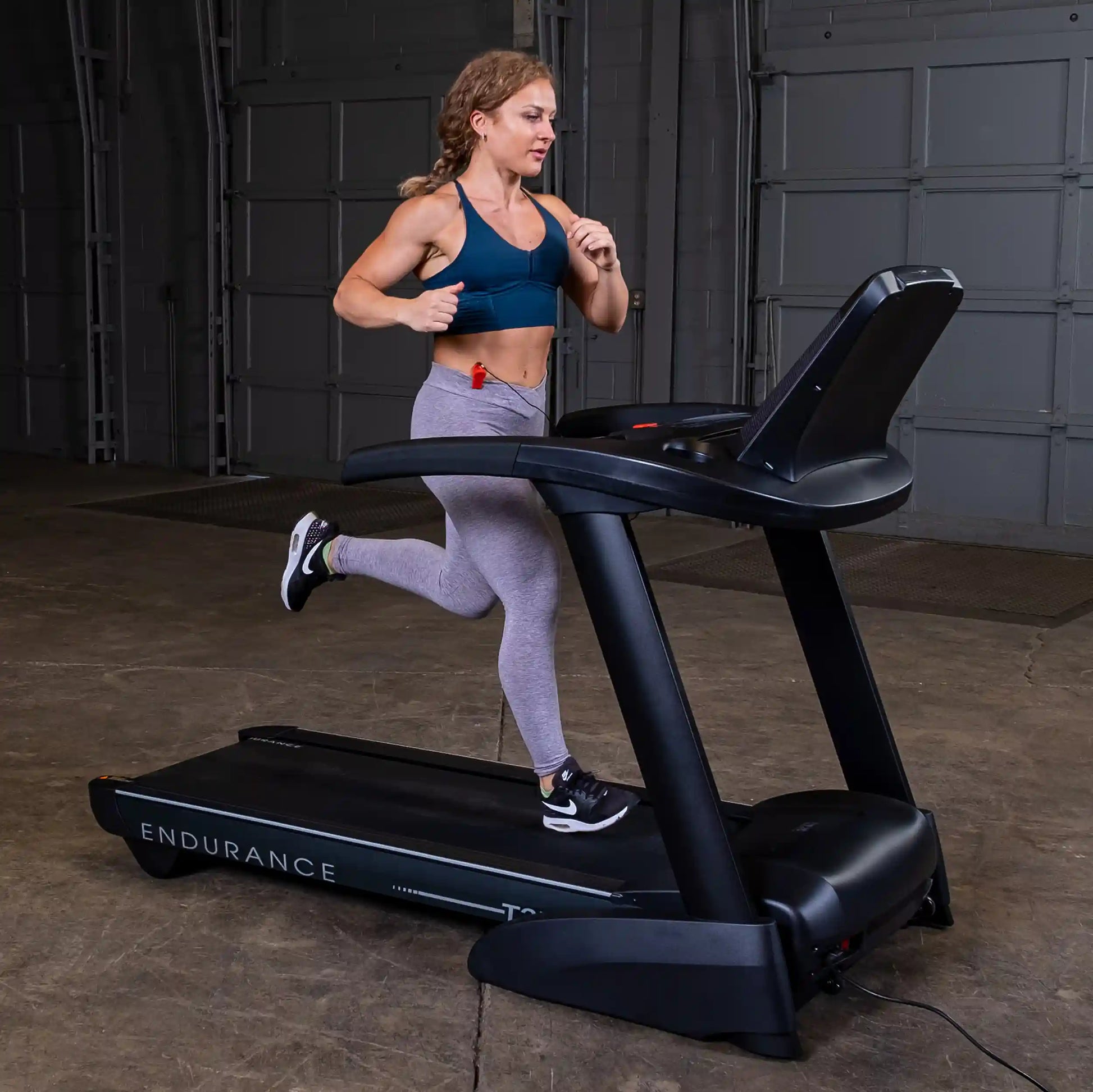Woman running on Endurance treadmill in a gym wearing gray leggings and a blue sports bra.