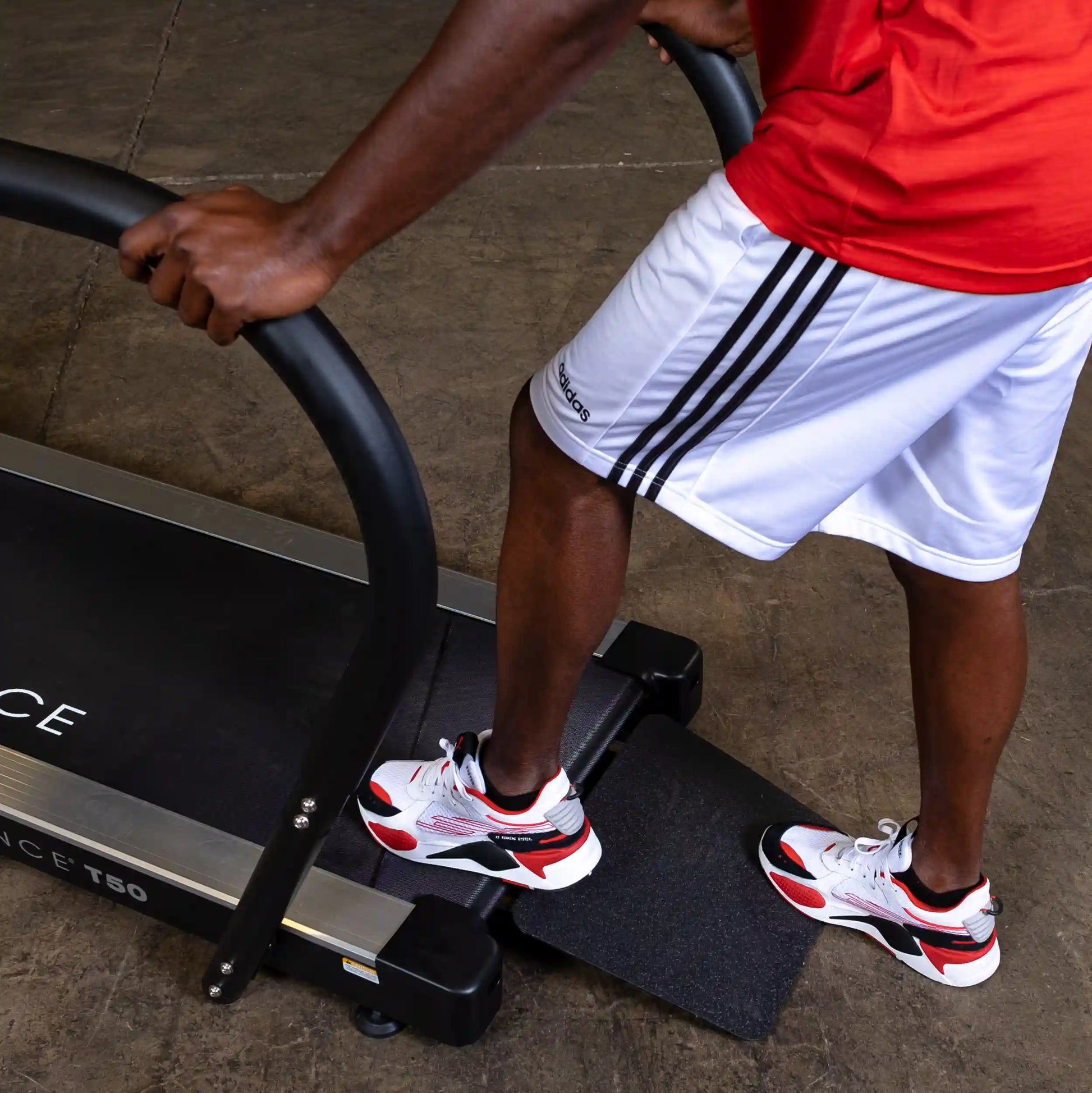 Man using curved treadmill, wearing Adidas shorts and red, black, and white sneakers.