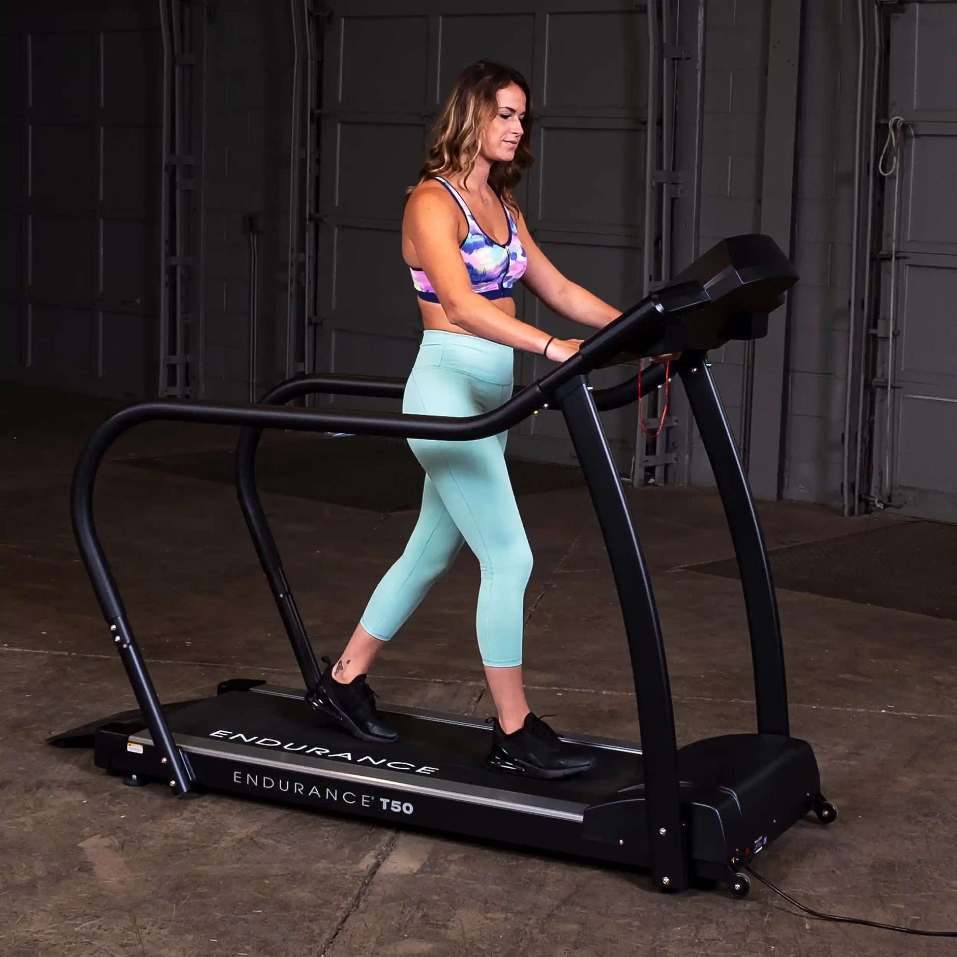 Woman walking on Endurance T50 treadmill in gym, wearing teal leggings and colorful sports bra.