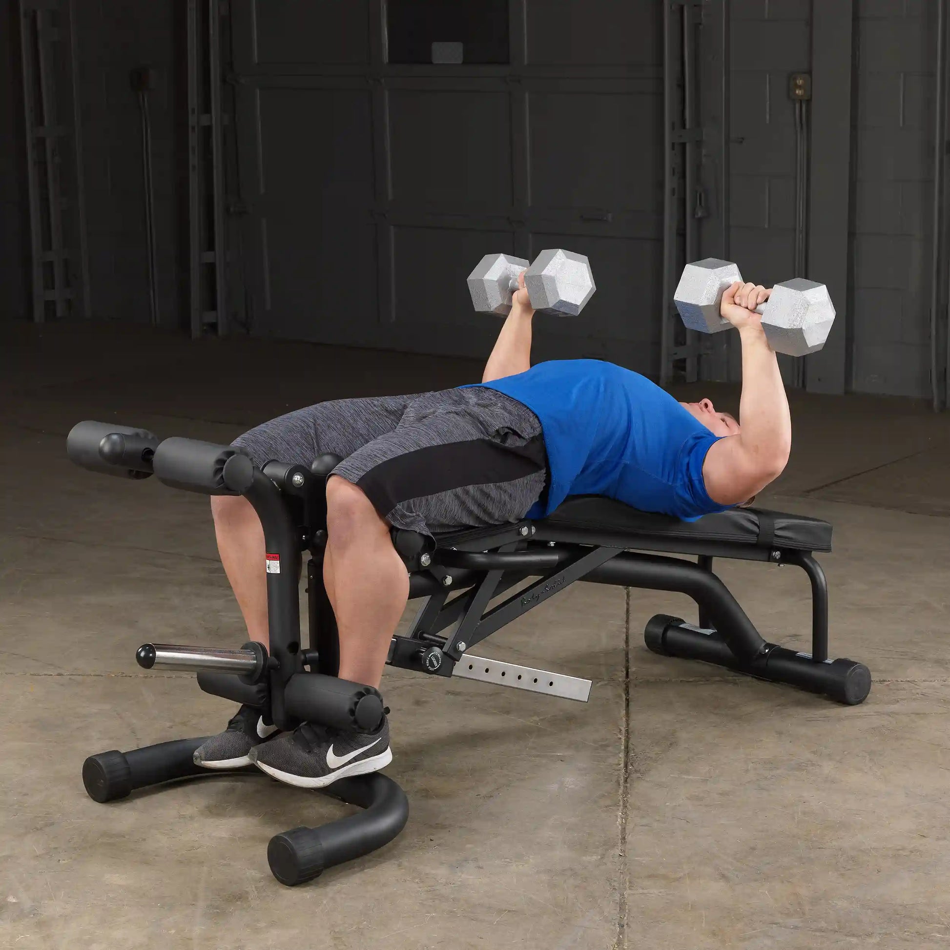 Man in blue shirt doing dumbbell press on black workout bench in gym.